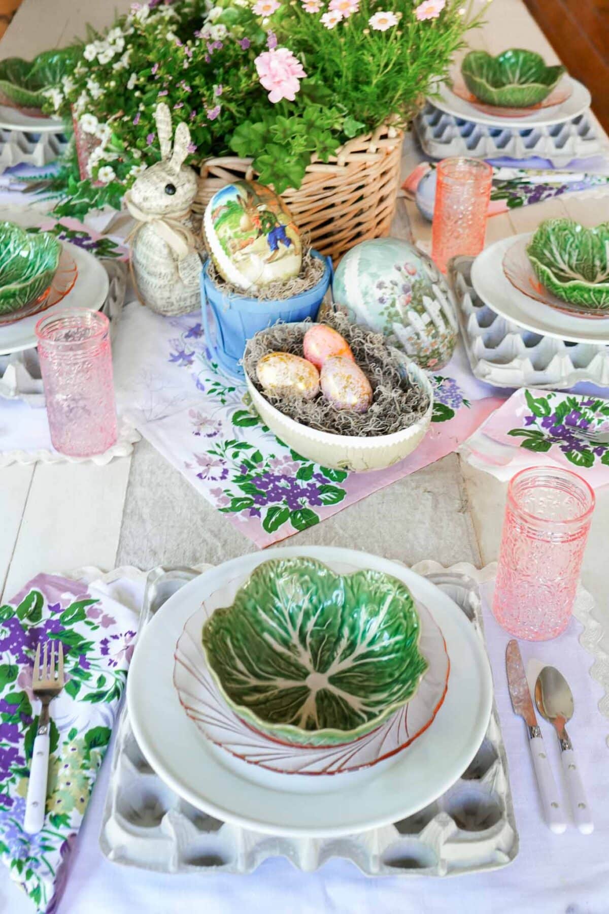 A spring-themed table setting with cabbage leaf plates on egg carton chargers, pink glasses, floral napkins, painted eggs, a bunny figurine, and a basket of flowers as the centerpiece—perfect for vintage Easter decorating.
