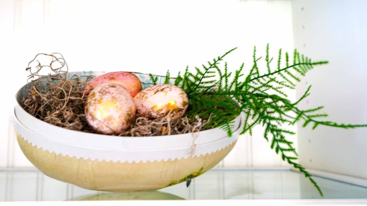 A decorative bowl filled with moss, three speckled pastel eggs, and a green fern branch, displayed on a glass shelf against a white background, captures the charm of vintage Easter decorating ideas.