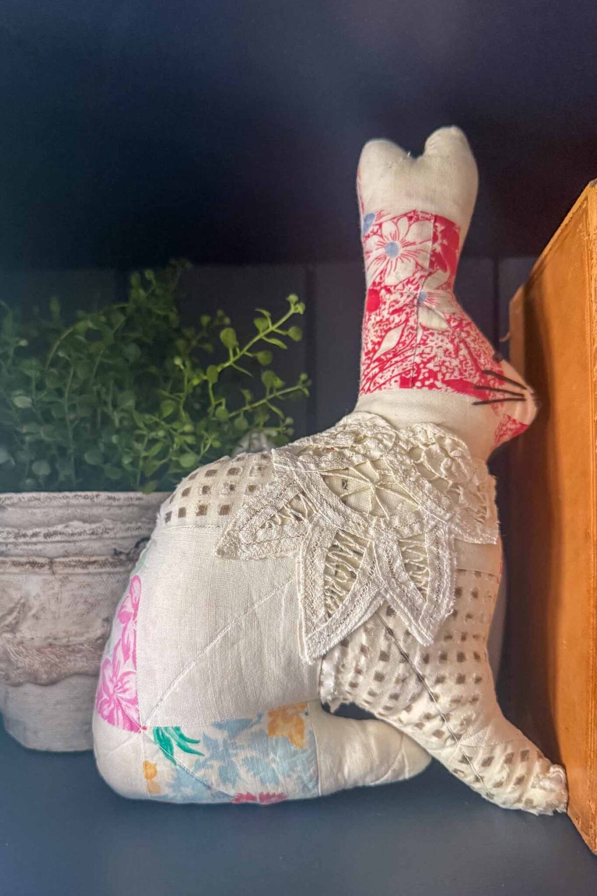 A handmade fabric bunny with patchwork and lace details sits on a shelf beside a small potted plant and an orange book, perfect for vintage Easter decorating ideas.