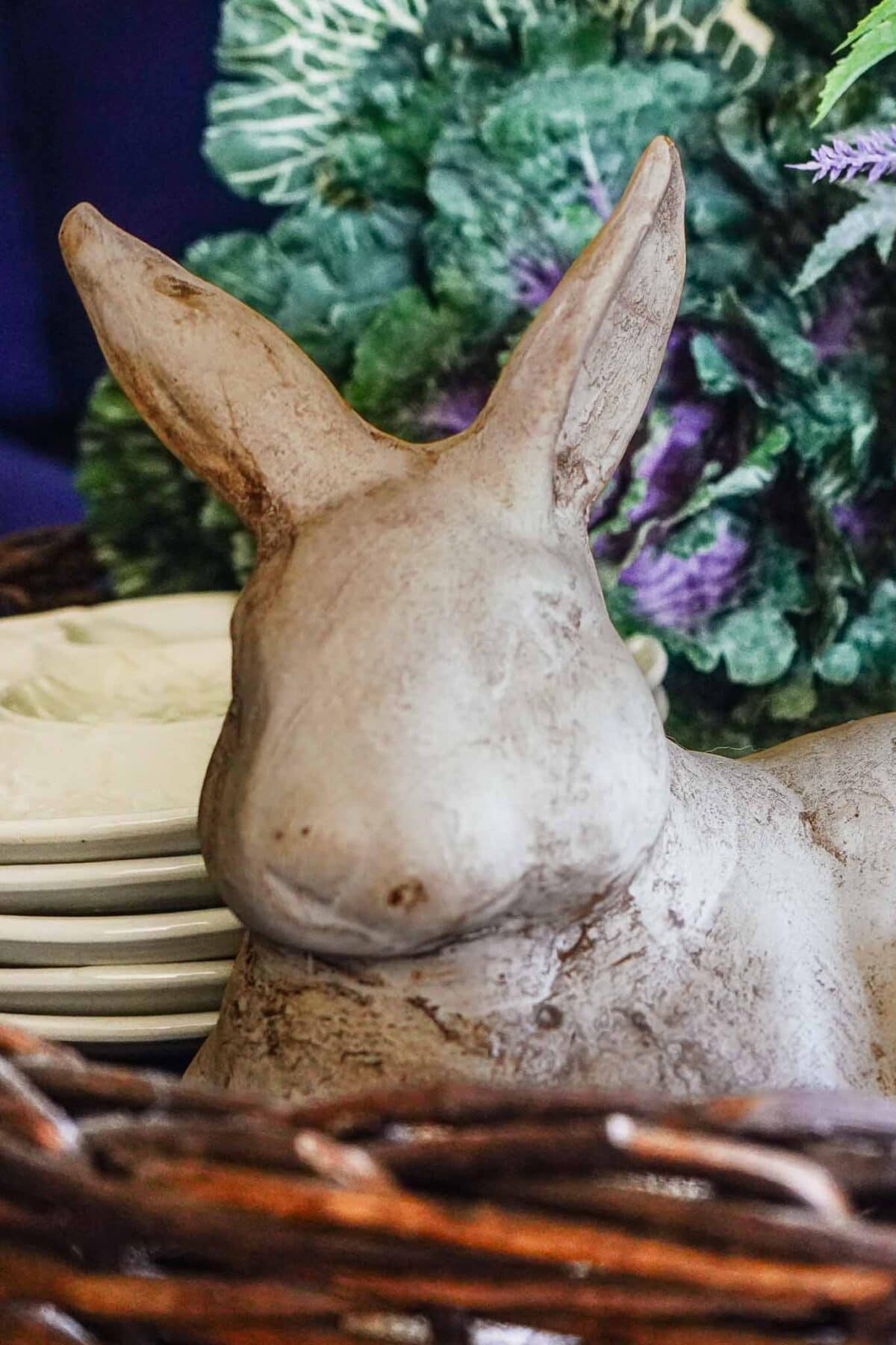 A close-up of a gray ceramic rabbit figurine placed in a wicker basket, offering charming vintage Easter decorating ideas, with stacked white plates and green foliage softly blurred in the background.