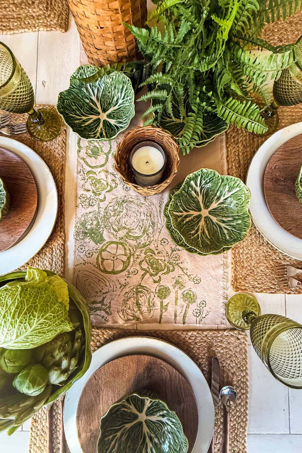 A dining table set with green leafy cabbage-shaped bowls, green glassware, woven placemats, white plates with wooden centers, a potted fern, a decorative candle in a basket, and napkins featuring vegetable block printing.