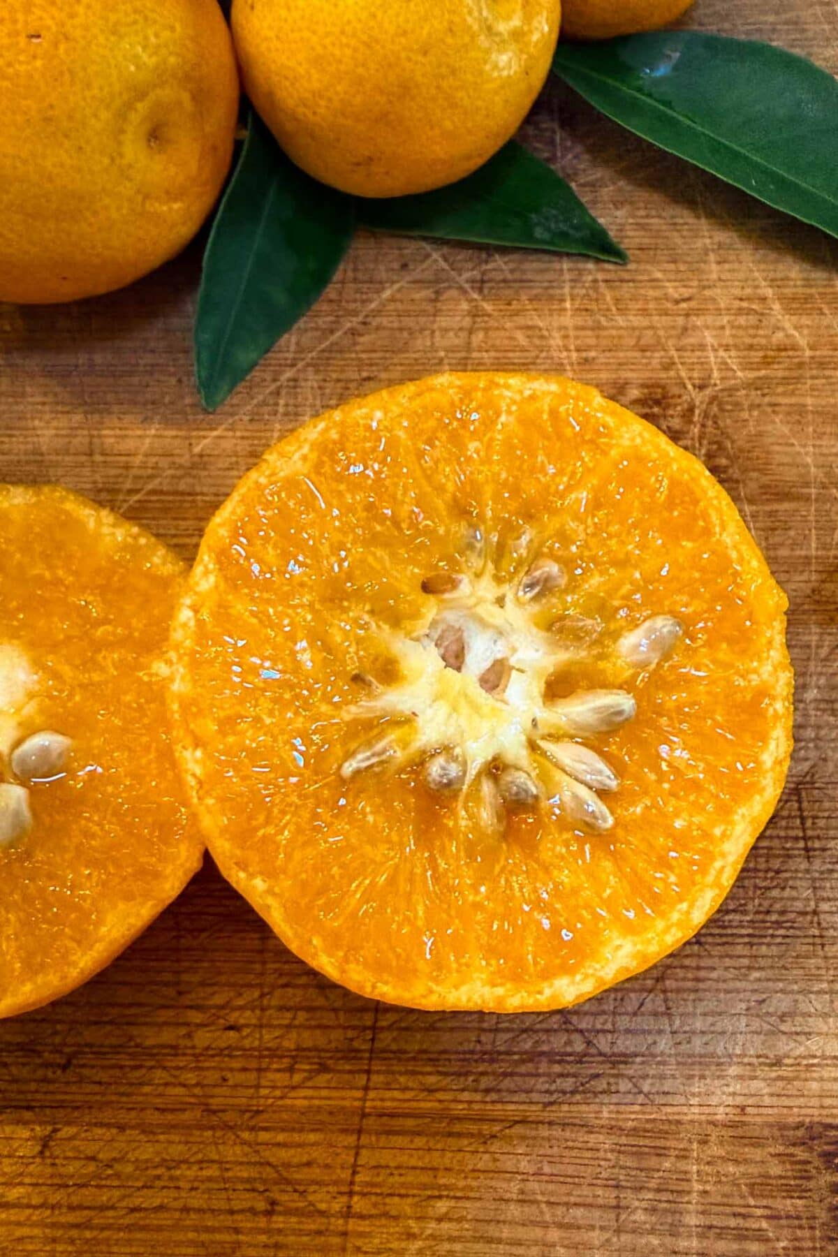 A close-up of a juicy orange cut in half on a wooden cutting board, with seeds visible inside. Whole oranges with green leaves and a splash of tangerine puree add color and freshness to the background.