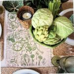 A dining table set with cabbage-themed decor, featuring a bowl of fresh green vegetables (cabbage, artichoke, celery), a woven candle holder, and a floral napkin adorned with cabbage vegetable block printing.