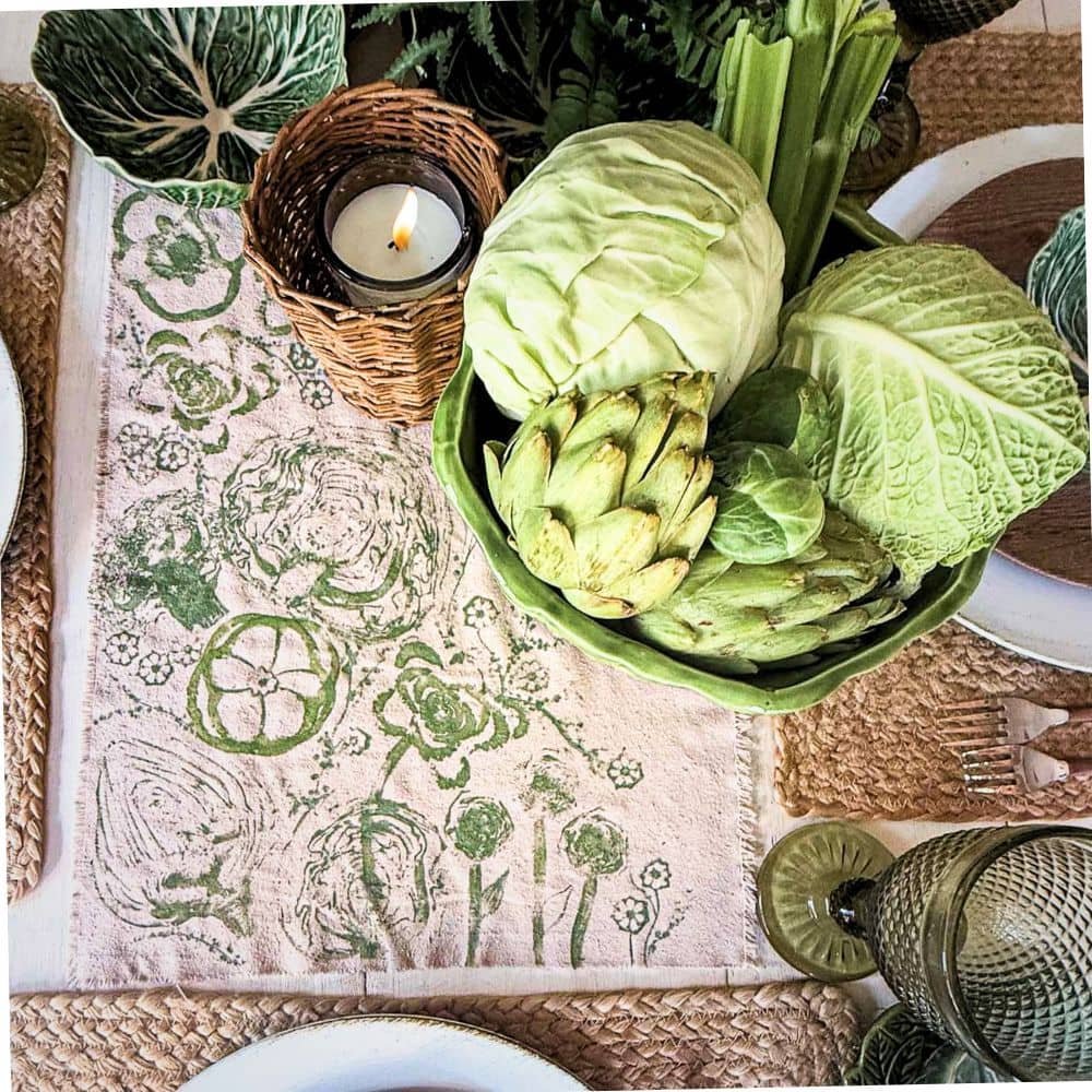 A dining table set with cabbage-themed decor, featuring a bowl of fresh green vegetables (cabbage, artichoke, celery), a woven candle holder, and a floral napkin adorned with cabbage vegetable block printing.