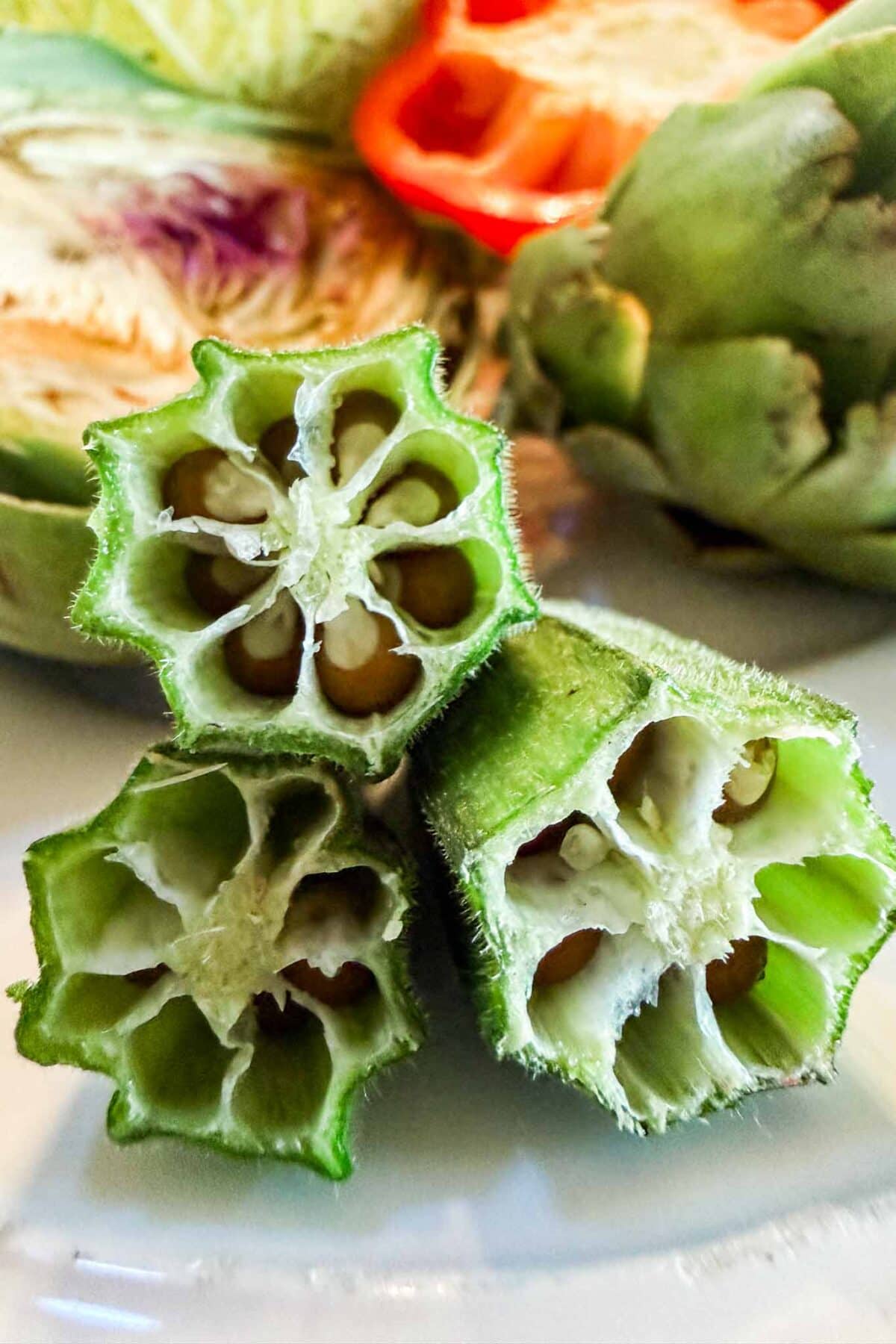 Close-up of three cross-sections of fresh green okra pods showing their star-shaped interiors, resembling patterns used in vegetable block printing, with artichokes and a sliced tomato blurred in the background.