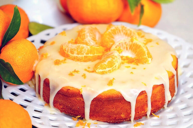 A round orange gluten-free tangerine cake with white icing drips, topped with mandarin orange segments and zest, sits on a white plate. Fresh mandarins with leaves are placed around the plate.
