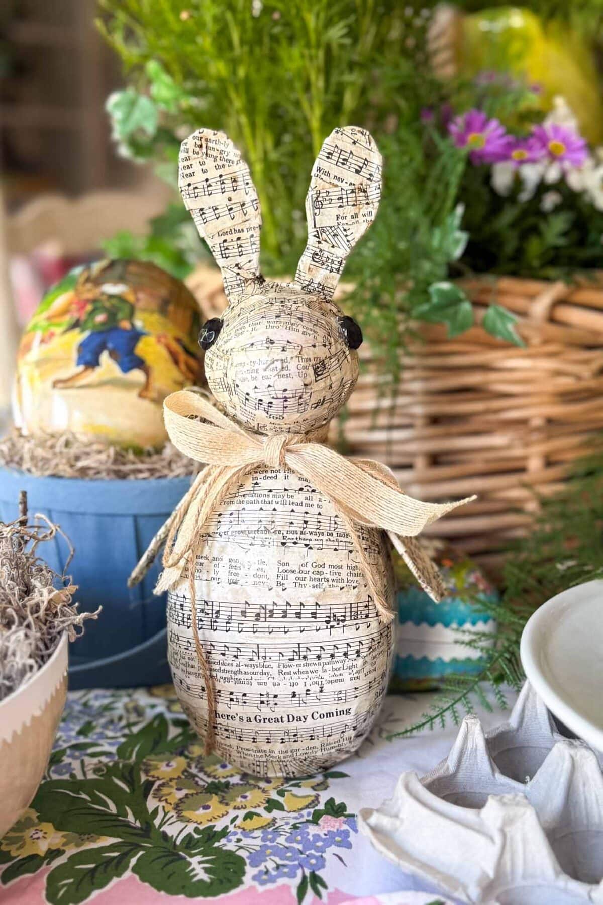 A decorative Paper Mache Easter Bunny covered in sheet music, wearing a burlap ribbon, sits on a floral tablecloth with greenery, a wicker basket, and colorful Easter eggs in the background.