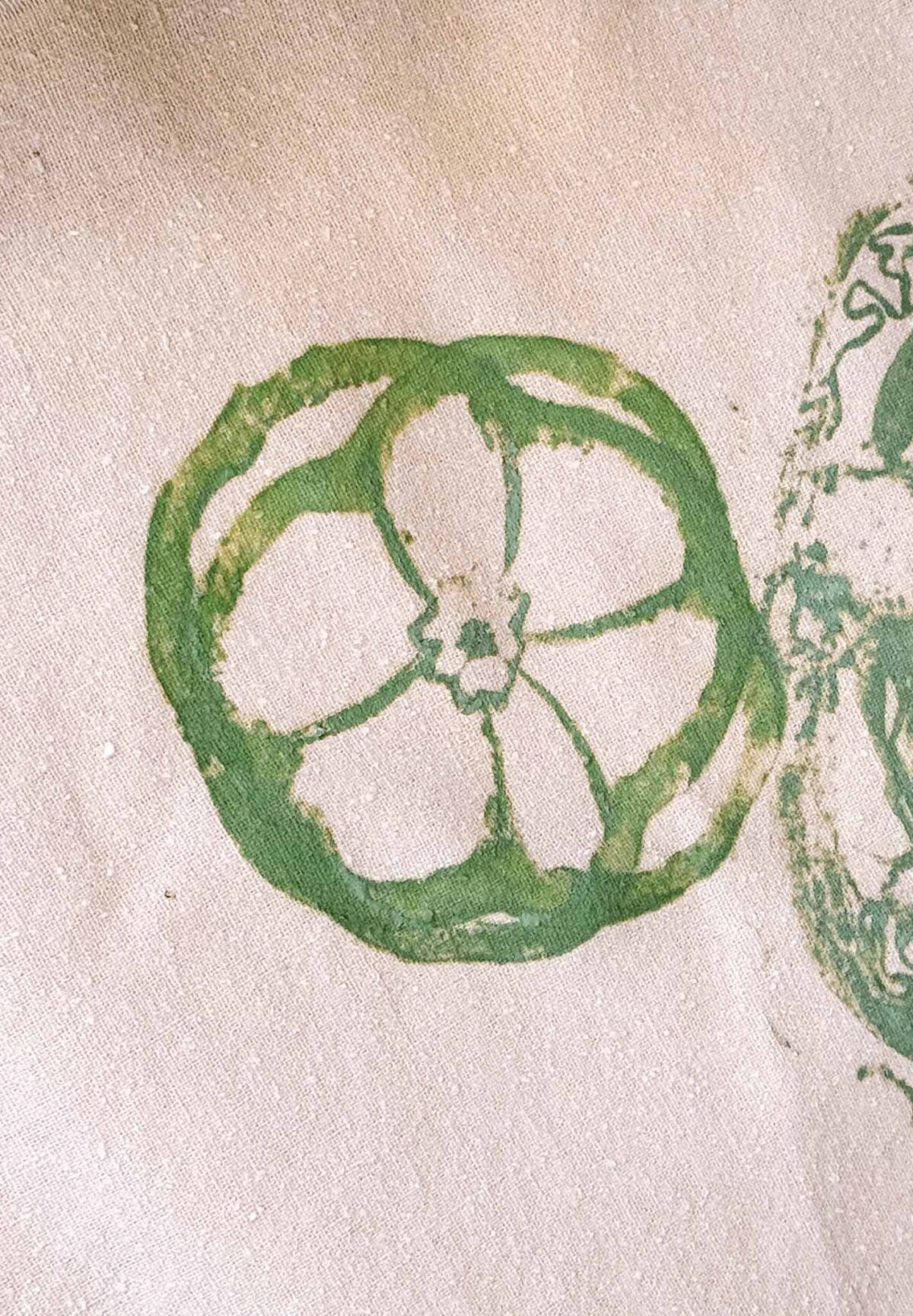 A green, circular stamp print created using vegetable block printing resembles the cross-section of a bell pepper on a light beige fabric background. The print shows five sections radiating from the center.