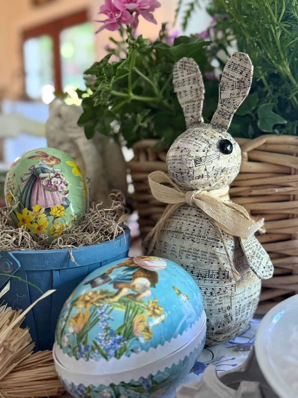 A decorative Paper Mache Easter Bunny made from sheet music sits beside painted Easter eggs and a blue pail of straw, surrounded by greenery and flowers in a wicker basket.