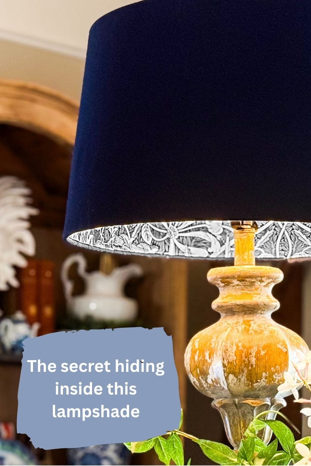 A decorative table lamp with a blue lampshade and wooden base stands in a cozy room. The underside features a black-and-white DIY lampshade lining with wallpaper. Overlaid text reads, “The secret hiding inside this lampshade.”.