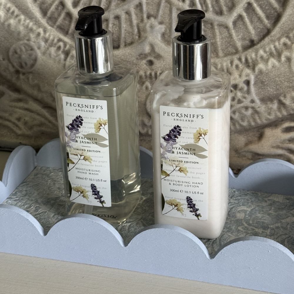 Two bottles of Pecksniff’s England Hyacinth & Jasmine hand wash and lotion with pump tops sit on a decorative tray with a scalloped edge, against a textured, patterned background.