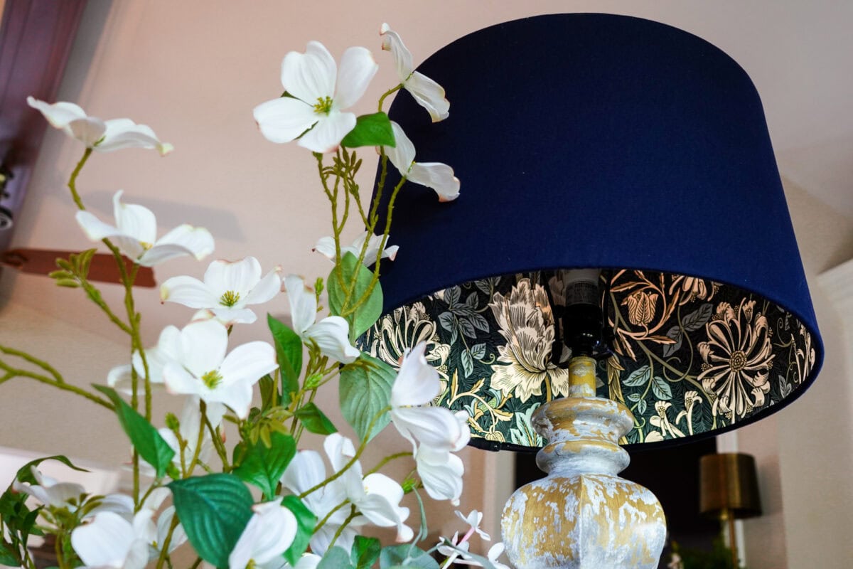 A decorative lamp with a dark blue shade featuring a gold floral pattern stands next to a bouquet of white flowers with green leaves. The background is softly lit and out of focus.