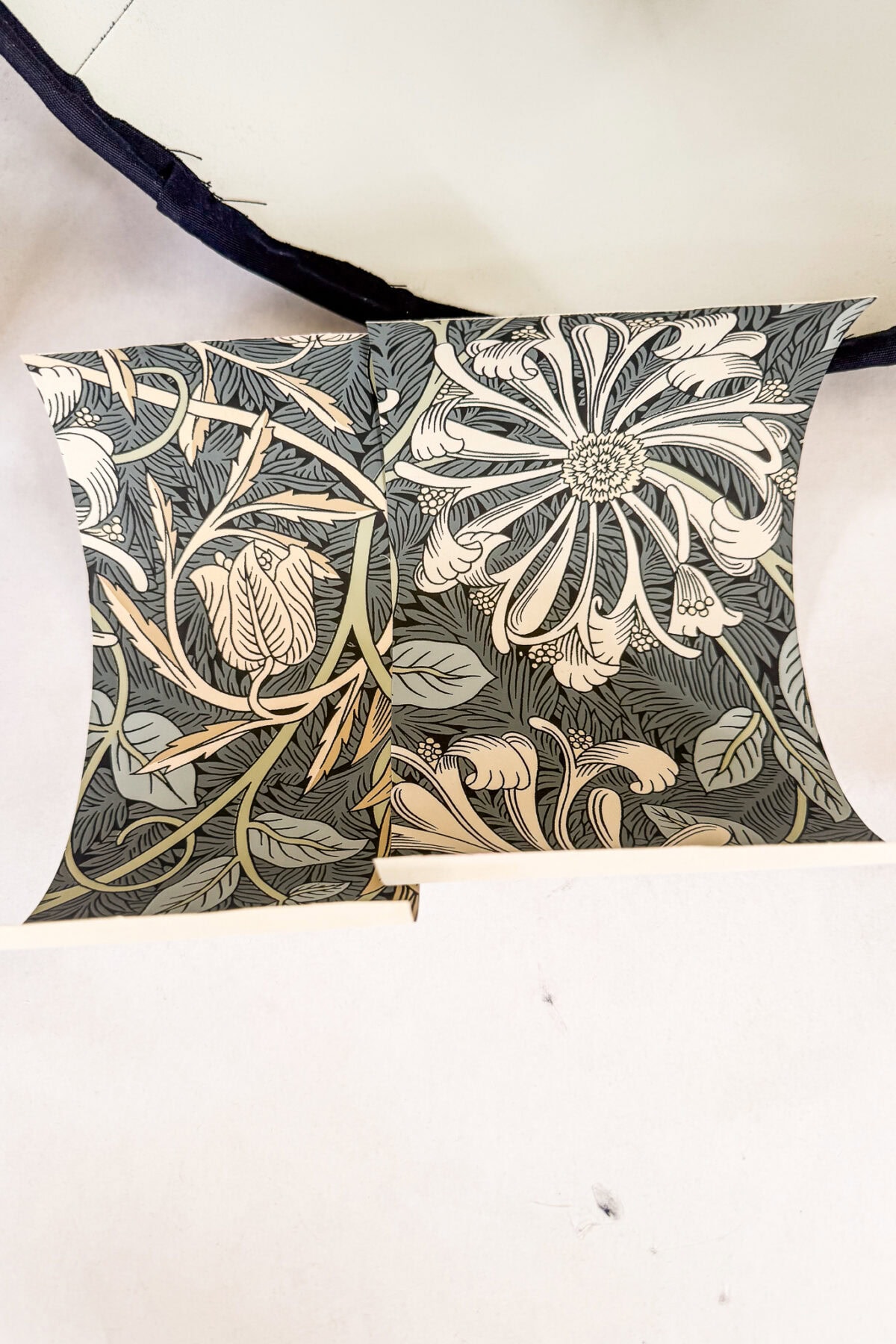 Two intricately patterned square pillows with botanical designs in beige and olive tones, featuring large flowers and leaves, resting on a light surface with a dark fabric edge visible at the top.