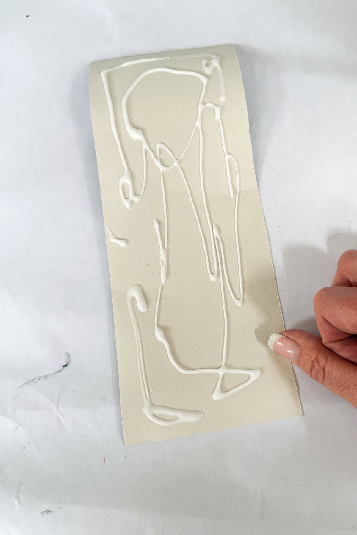 A hand holding a rectangular piece of off-white paper with irregular lines of white glue squirted across its surface, placed on a white background.