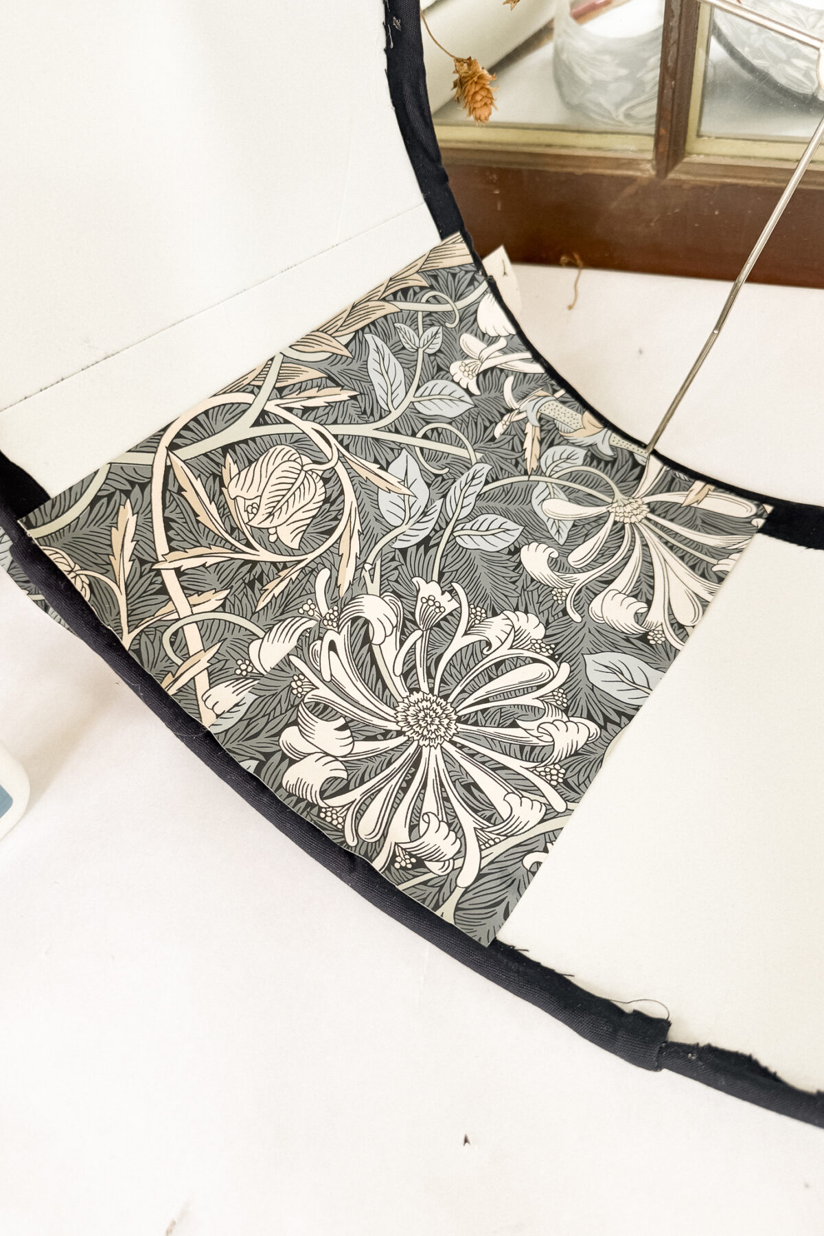 A close-up of a floral-patterned paper or fabric being applied to the inside of a curved white surface, with a window and some dried plants in the background.