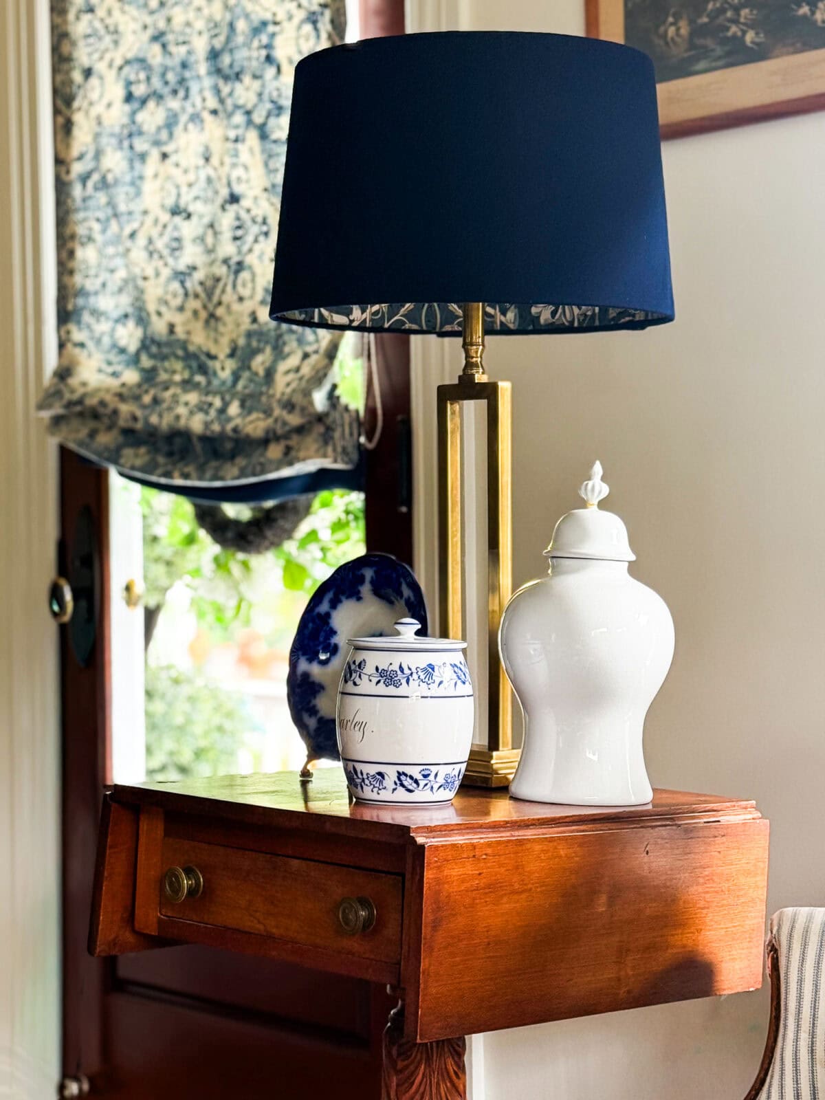 A wooden side table with two drawers holds a gold and blue lamp, a white decorative jar, and two blue and white porcelain plates in front of a window with a patterned shade and leafy greenery outside.