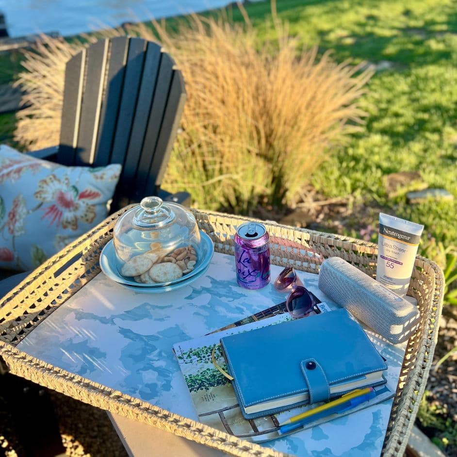 A wicker tray on an outdoor table holds cookies under a glass dome, a can of LaCroix, a tube of sunscreen, sunglasses, a rolled-up towel, a blue planner, pens, and magazines. A lawn chair and grass are in the background.
