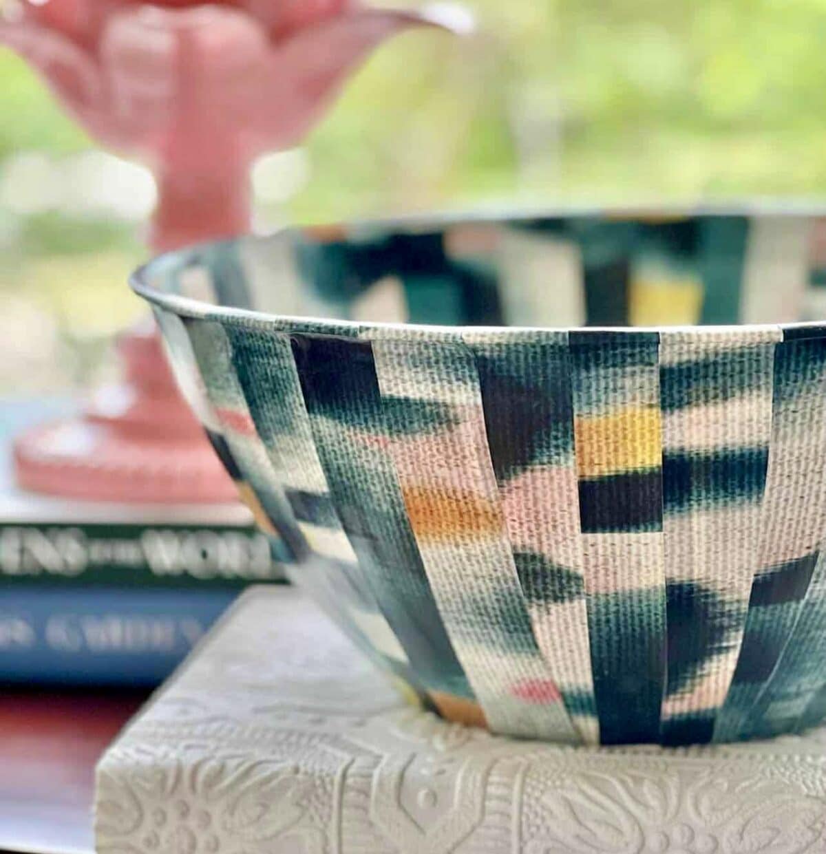 A colorful, patterned bowl with a geometric design sits on an embossed white surface, with blurred books and a pink decorative object in the background.