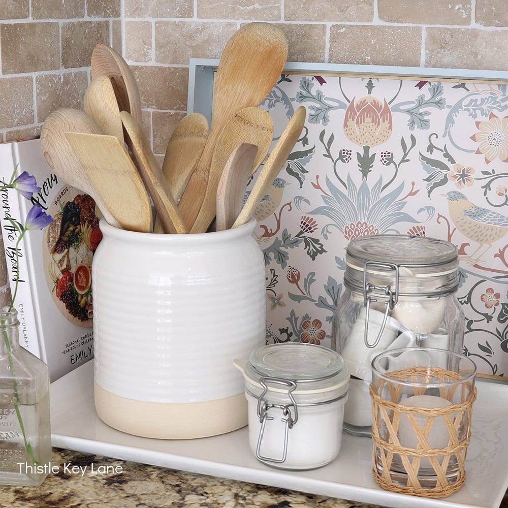 A white ceramic jar holds various wooden kitchen utensils beside glass jars of sugar and salt on a white tray. A decorative floral recipe book and a candle in a woven holder complete the cozy kitchen scene.