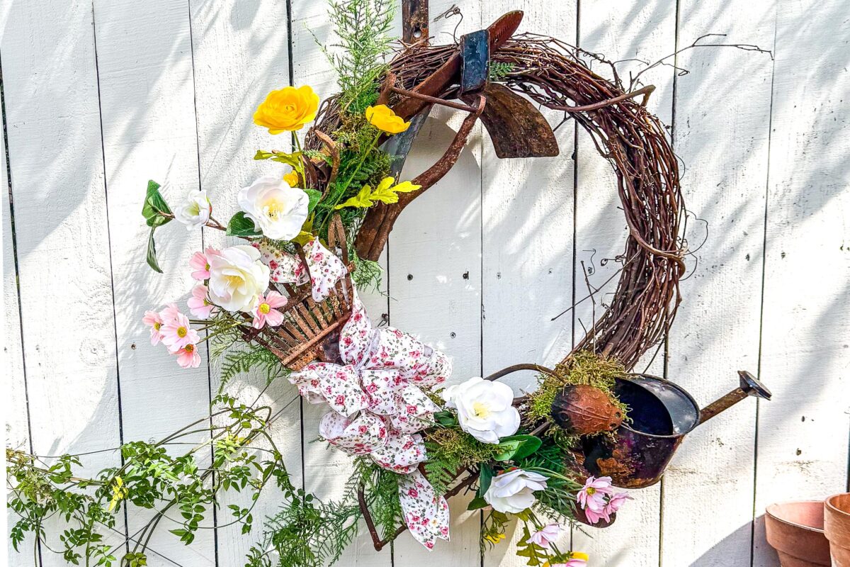 A rustic wreath made of twigs hangs on a white wooden fence, decorated with artificial flowers, a small watering can, a trowel, greenery, and a pink floral ribbon. Sunlight casts soft shadows over the scene.