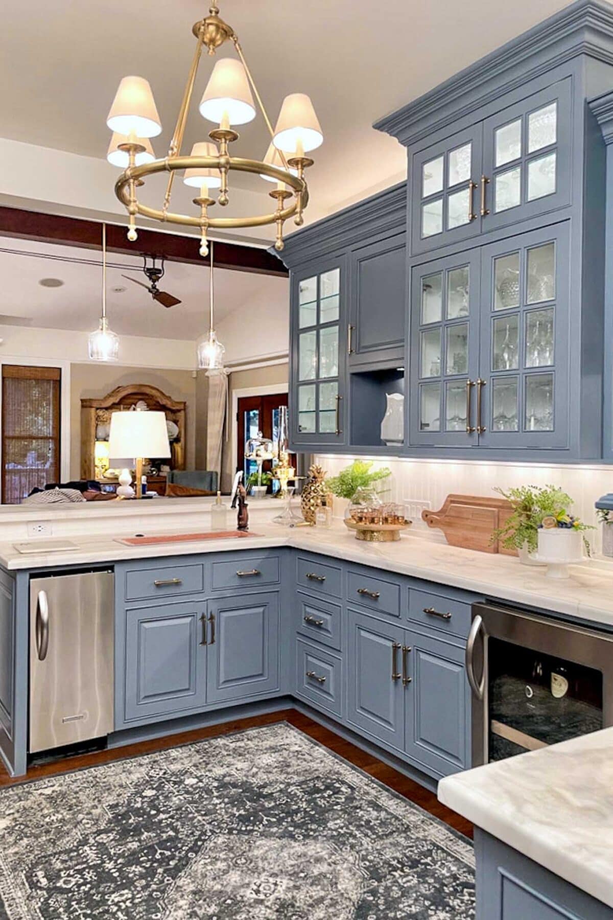 A modern kitchen with blue cabinets, marble countertops, glass-front upper cabinets, stainless steel appliances, pendant lights, and a patterned rug on a dark wood floor. Plants and decorative items are on the counters.
