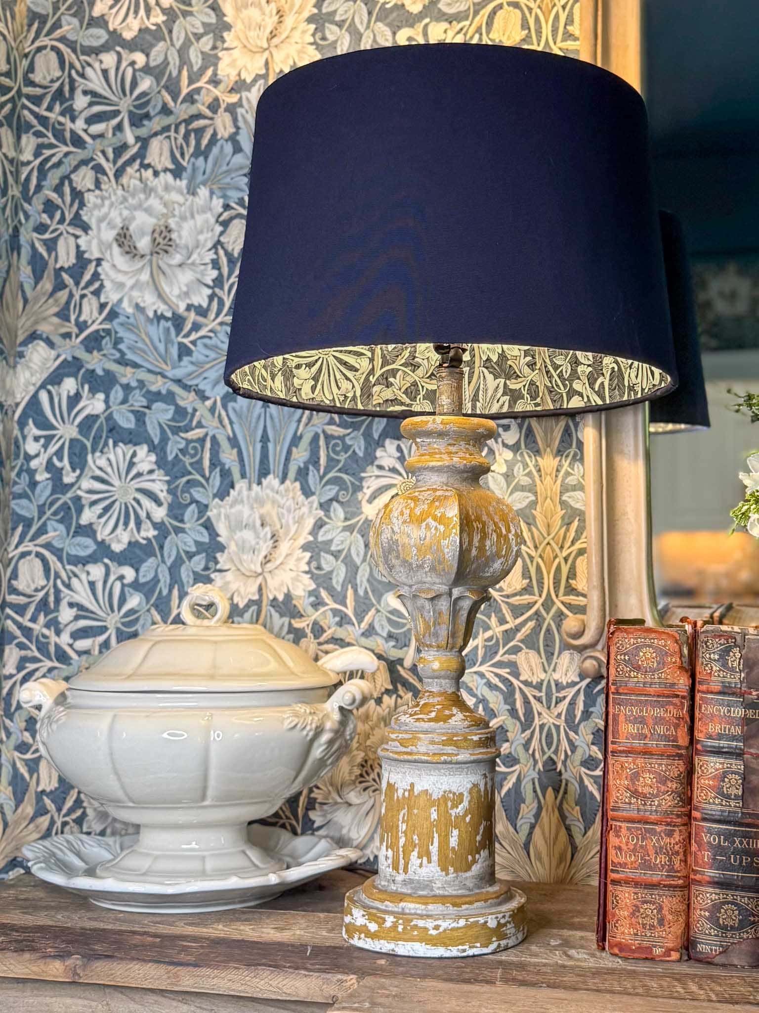 A rustic yellow lamp with a DIY lampshade lining with wallpaper sits on a wooden table next to vintage books and a white ceramic tureen, against a floral-patterned wallpaper background.