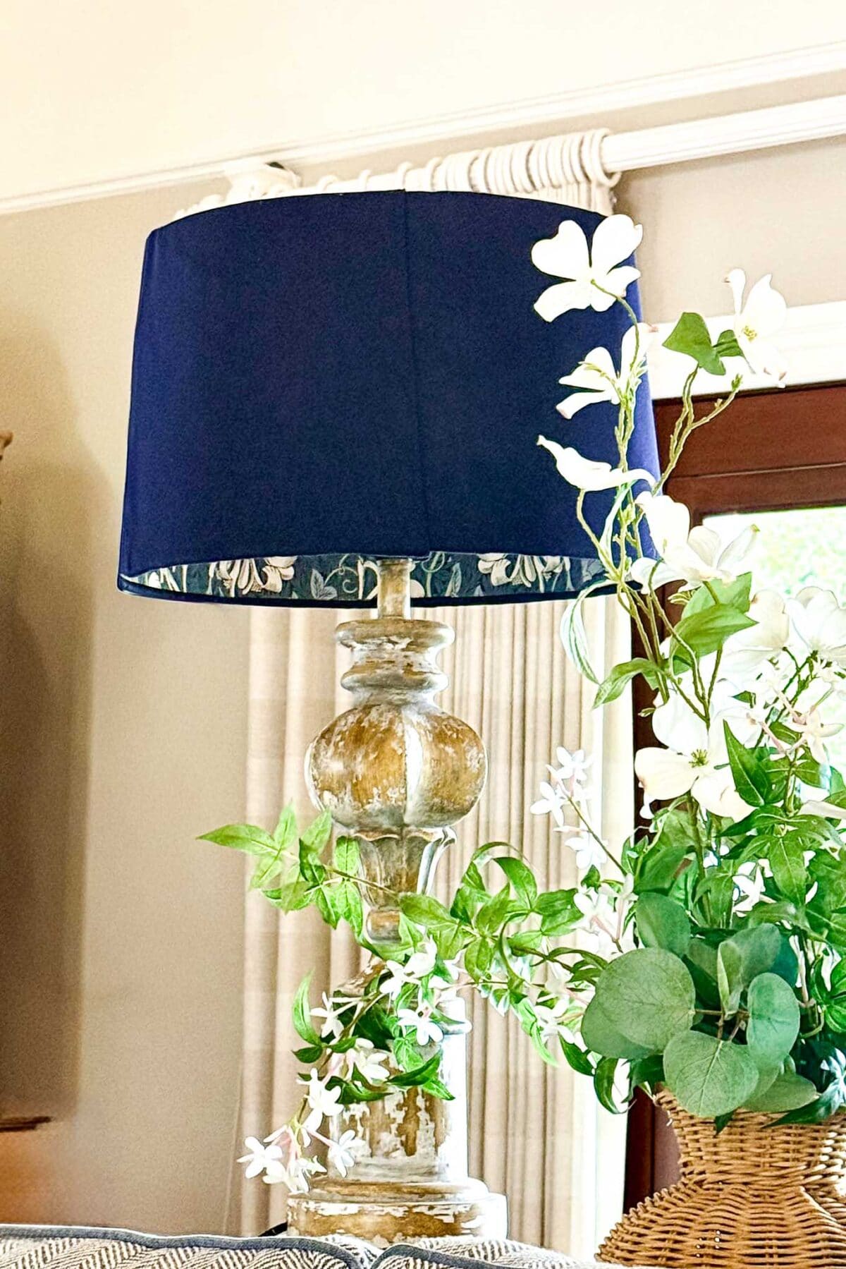 A vintage-style table lamp with a blue DIY lampshade lining with wallpaper sits next to a green plant with white flowers. The scene is bright, with curtains and a window in the background.