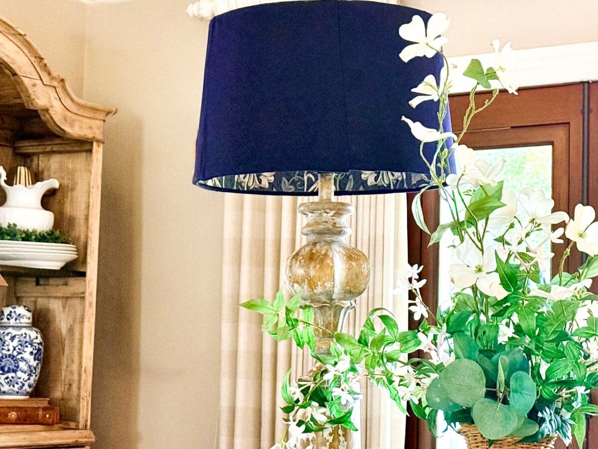 A decorative lamp with a dark blue shade featuring DIY lampshade lining with wallpaper sits next to a bouquet of white flowers and green leaves in a well-lit room with beige walls and wooden furniture.