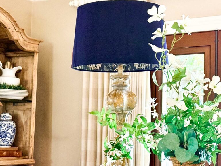 A decorative lamp with a dark blue shade featuring DIY lampshade lining with wallpaper sits next to a bouquet of white flowers and green leaves in a well-lit room with beige walls and wooden furniture.