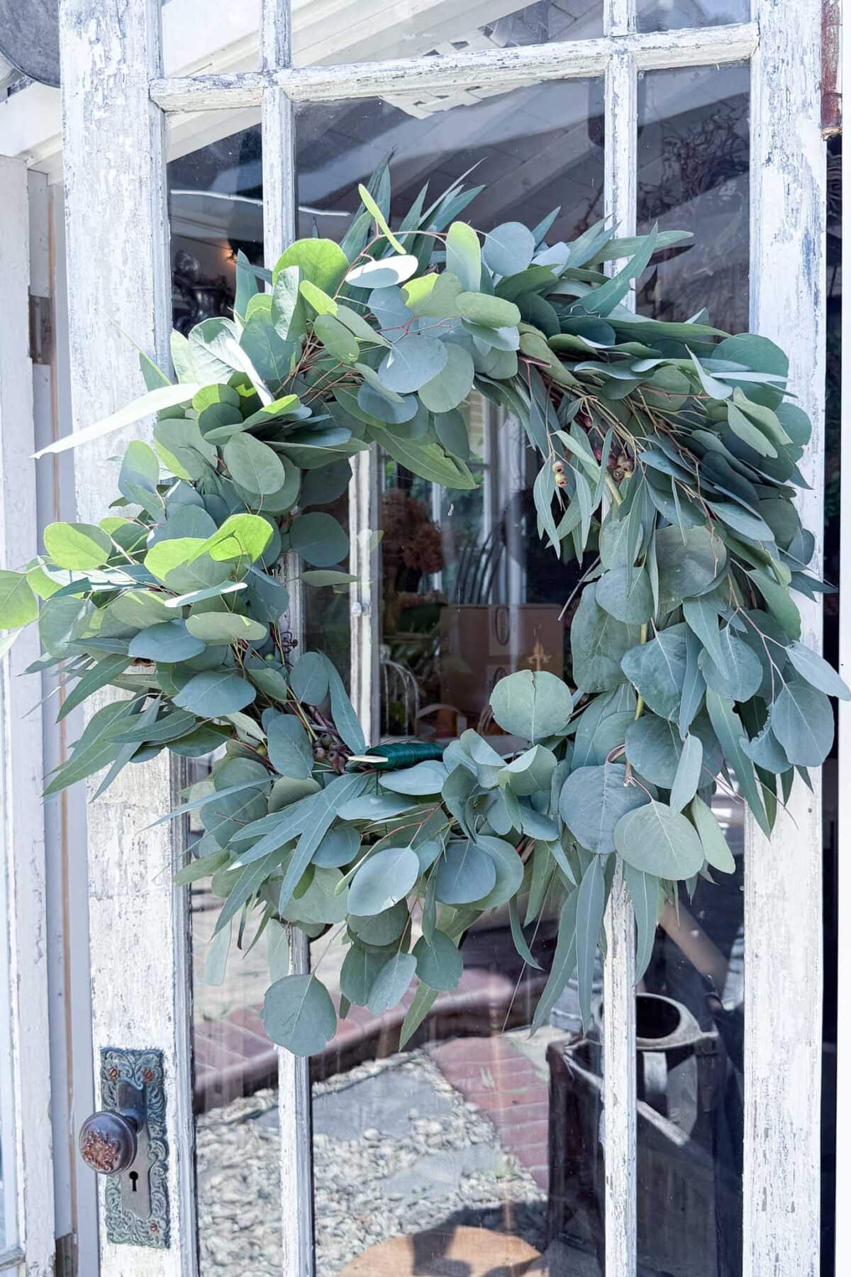 A lush eucalyptus wreath hangs on a weathered white wooden door with glass panes.