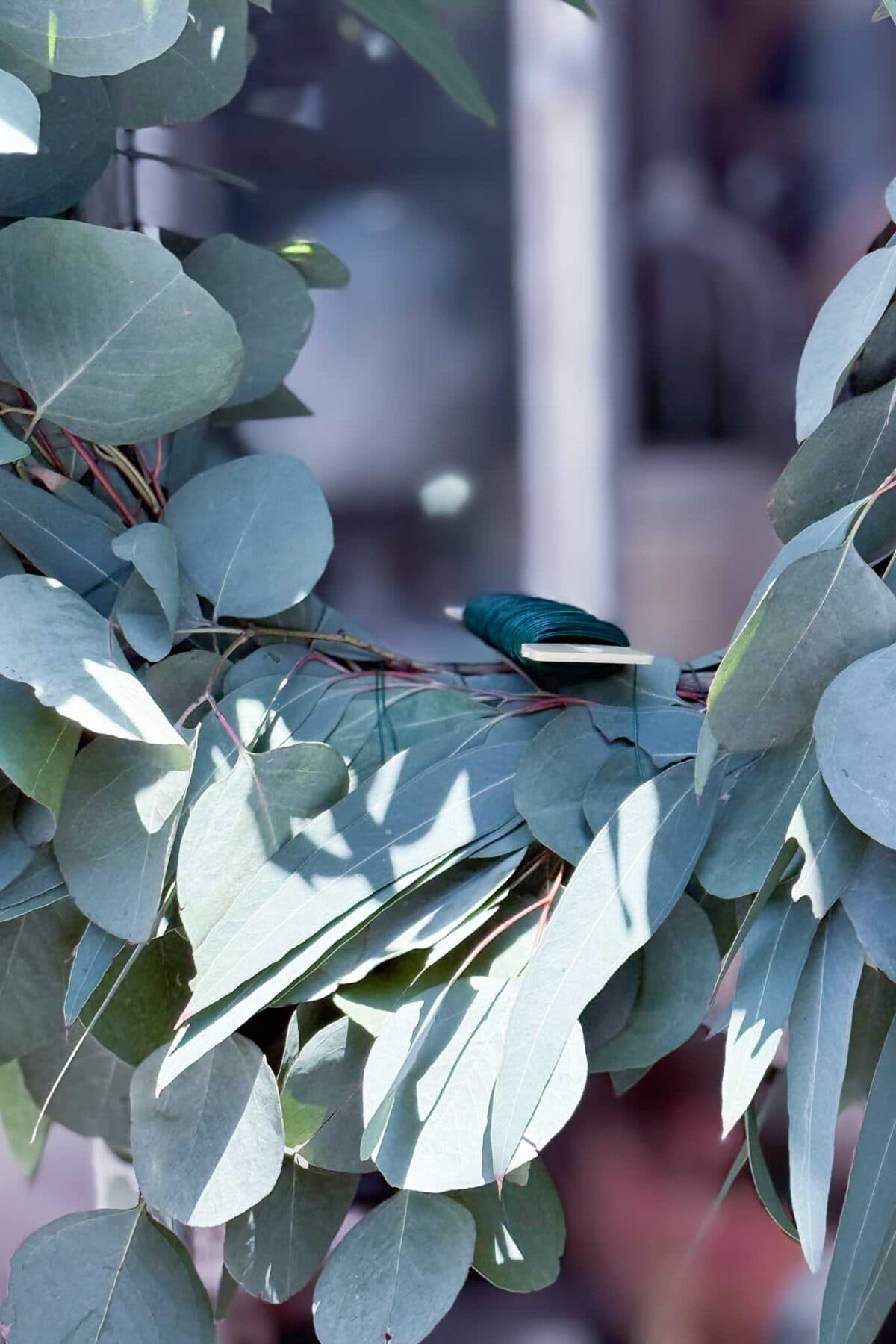 A spool of green floral wire rests on a wreath of eucalyptus leaves, partially obscuring the soft focus background.