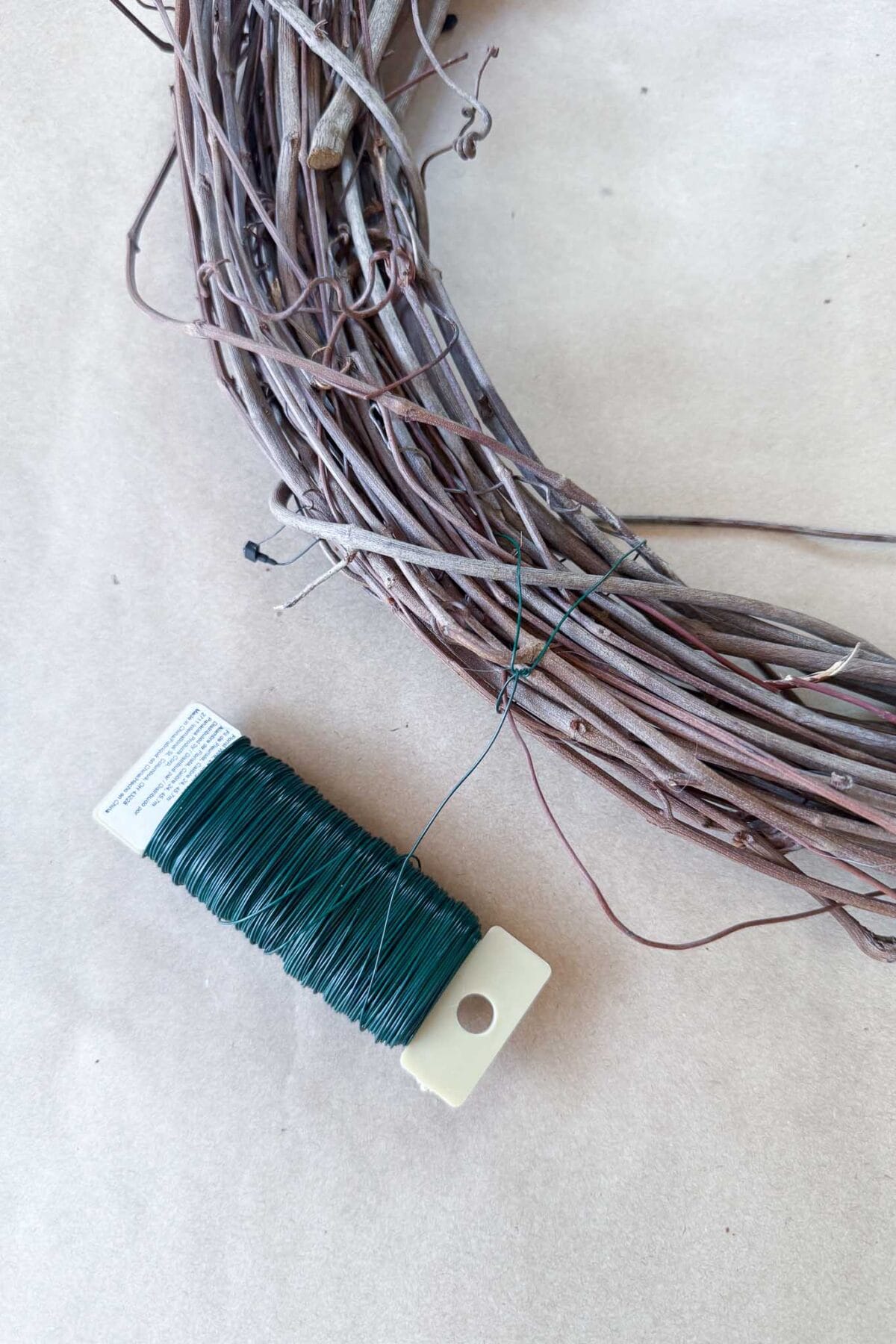 A spool of green floral wire lies next to a twig wreath base on a tan background.