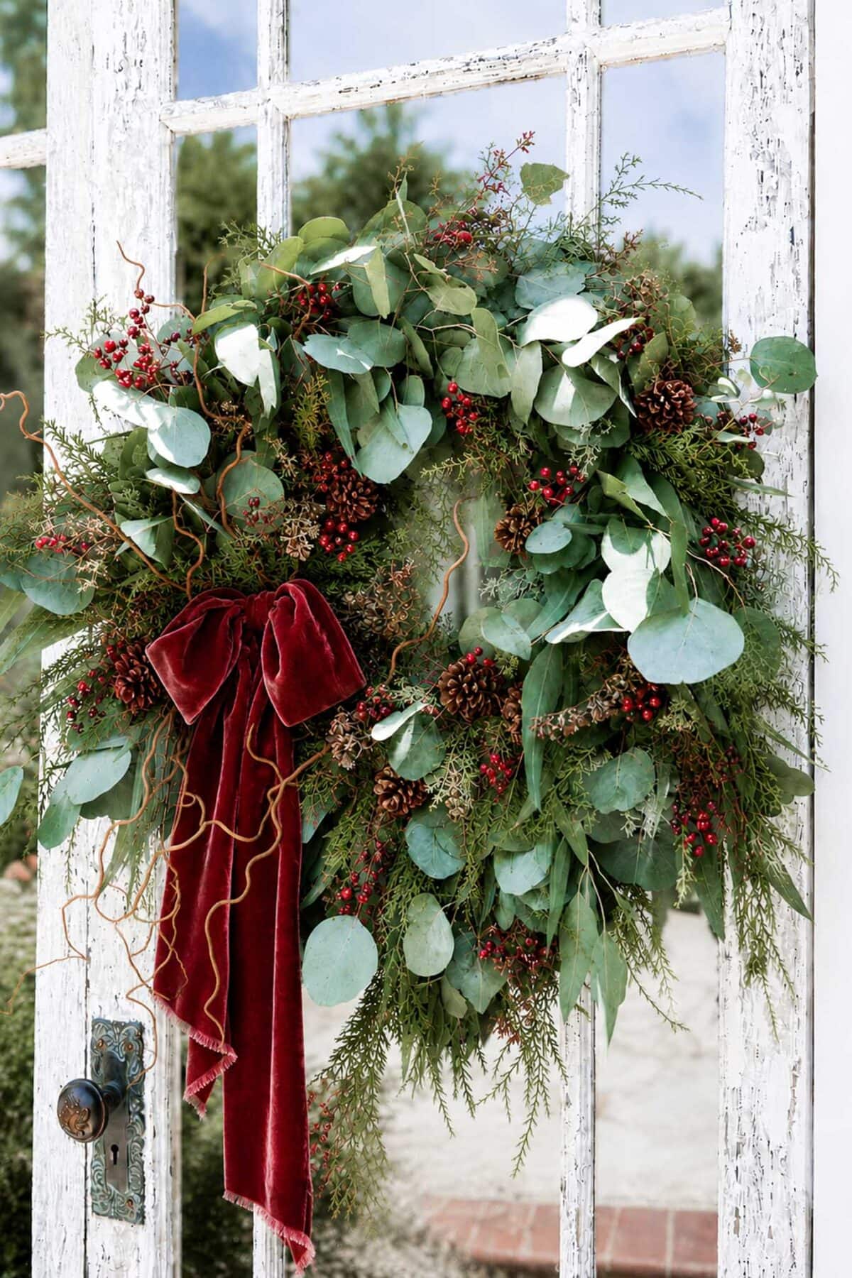A rustic wreath made of eucalyptus, pinecones, red berries, and greenery hangs on a weathered white door, adorned with a deep red velvet ribbon.