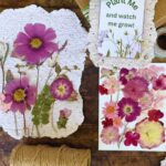 Handmade seed paper decorated with pressed pink and purple flowers lies on a table with twine, floral cutouts, and a “Plant Me and watch me grow!” tag attached to a gift box—perfect for DIY seed paper handmade cards or unique stationery.