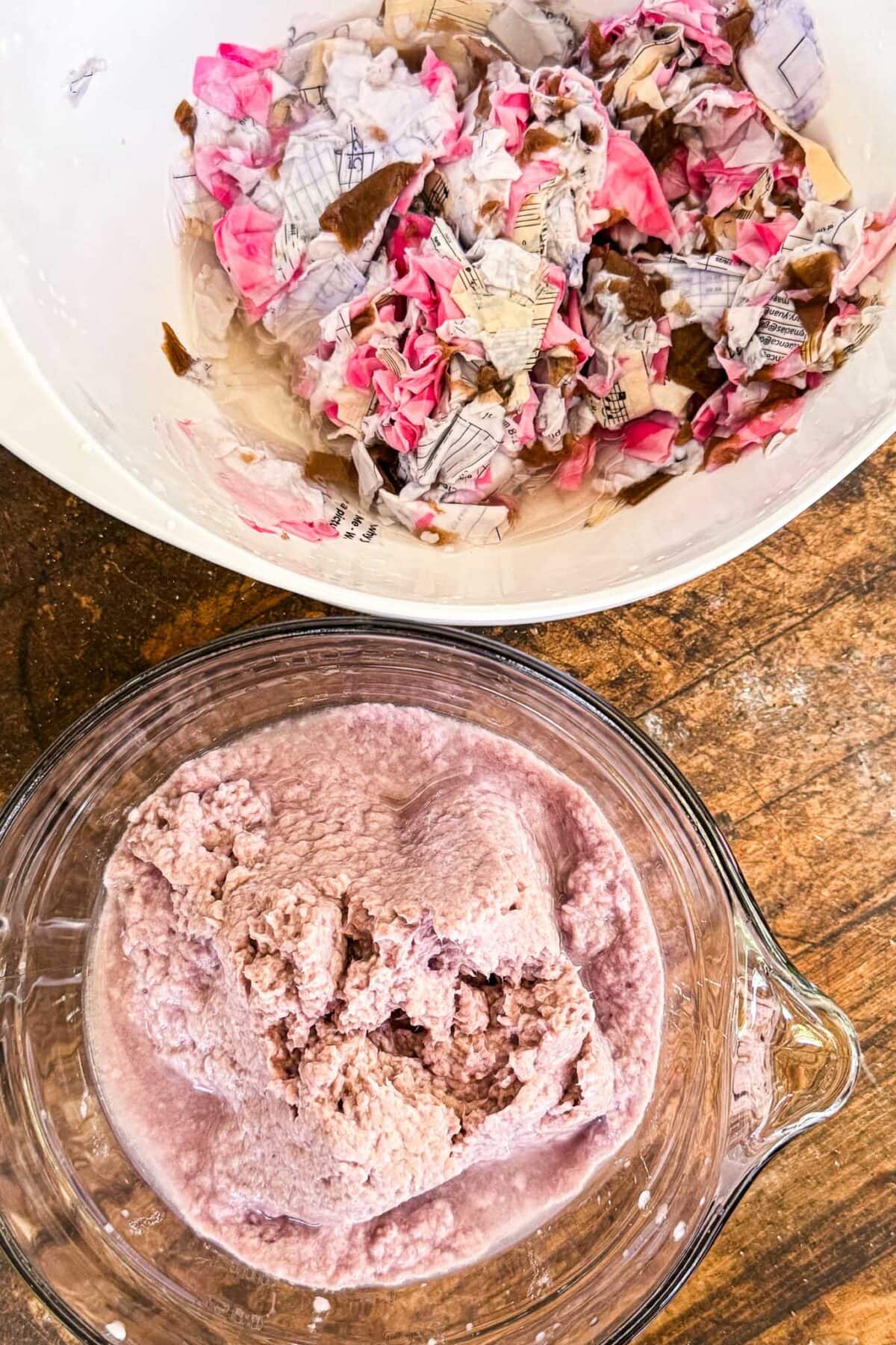Two bowls on a wooden surface: one with torn, wet pieces of paper in shades of pink, white, and brown; the other with blended, pulpy, light pink paper mixture.
