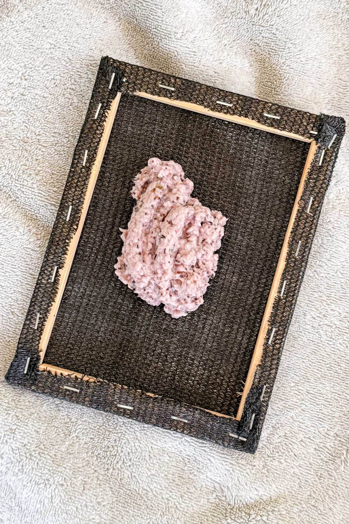 A mound of pinkish pulp sits on a black mesh screen framed with wood, placed on a light-colored textured surface.