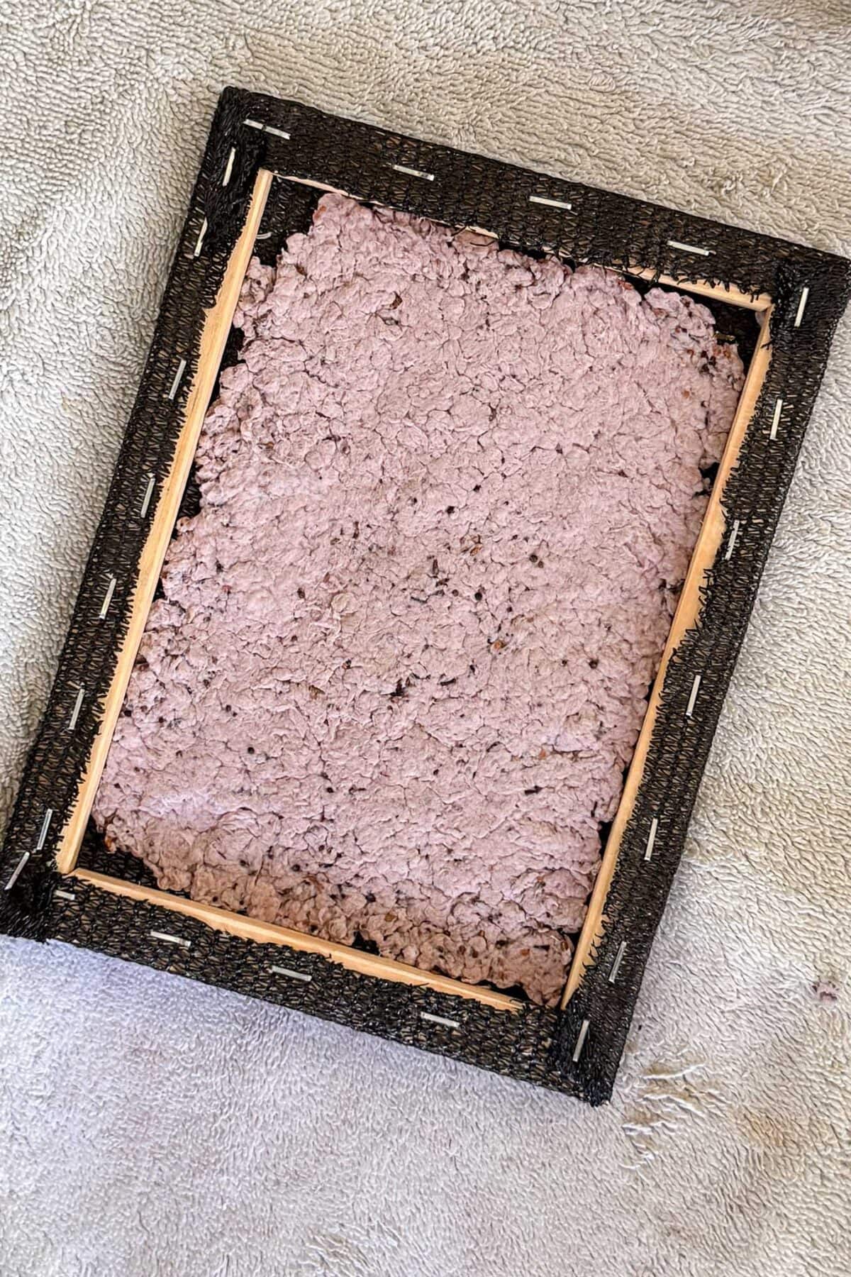 A wooden frame with a backing of torn black material is filled with a layer of rough, handmade recycled paper pulp in a light purple color, placed on a textured beige fabric surface.