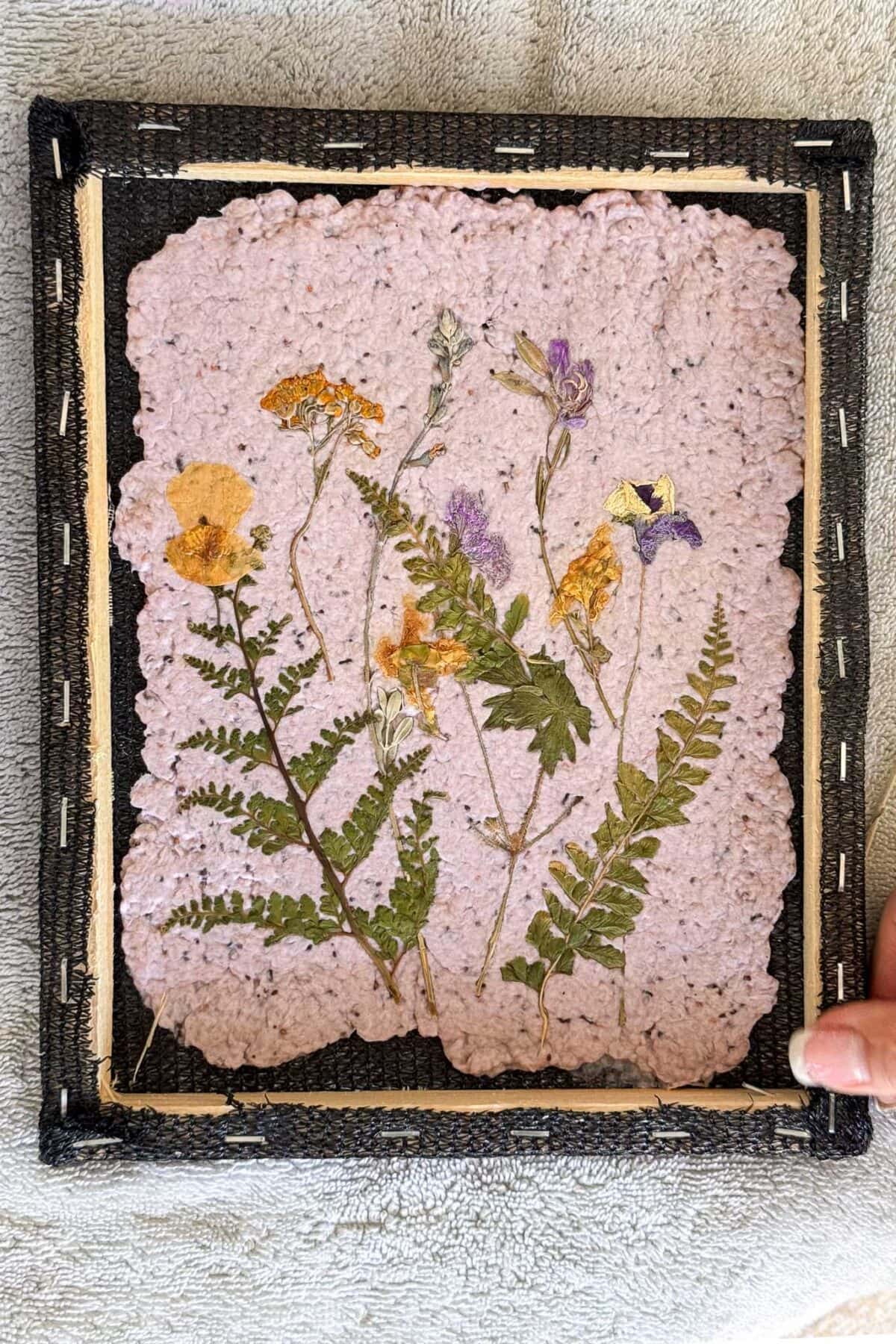 A hand holds a framed piece of handmade paper with pressed yellow, orange, purple flowers, and green ferns. The paper has rough edges and is mounted on a black textured background with visible staples along the border.