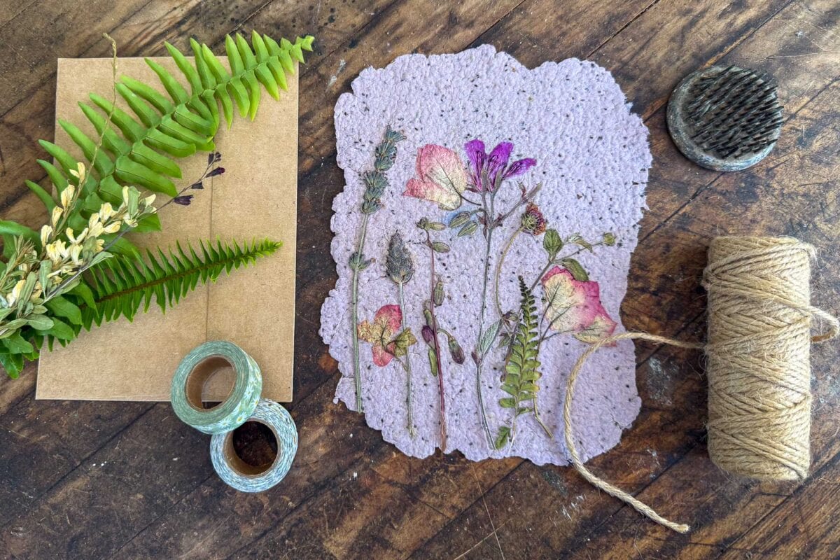A handmade card with pressed flowers lies on a wooden table beside fern leaves, an envelope, two rolls of washi tape, twine, and a metal flower frog.