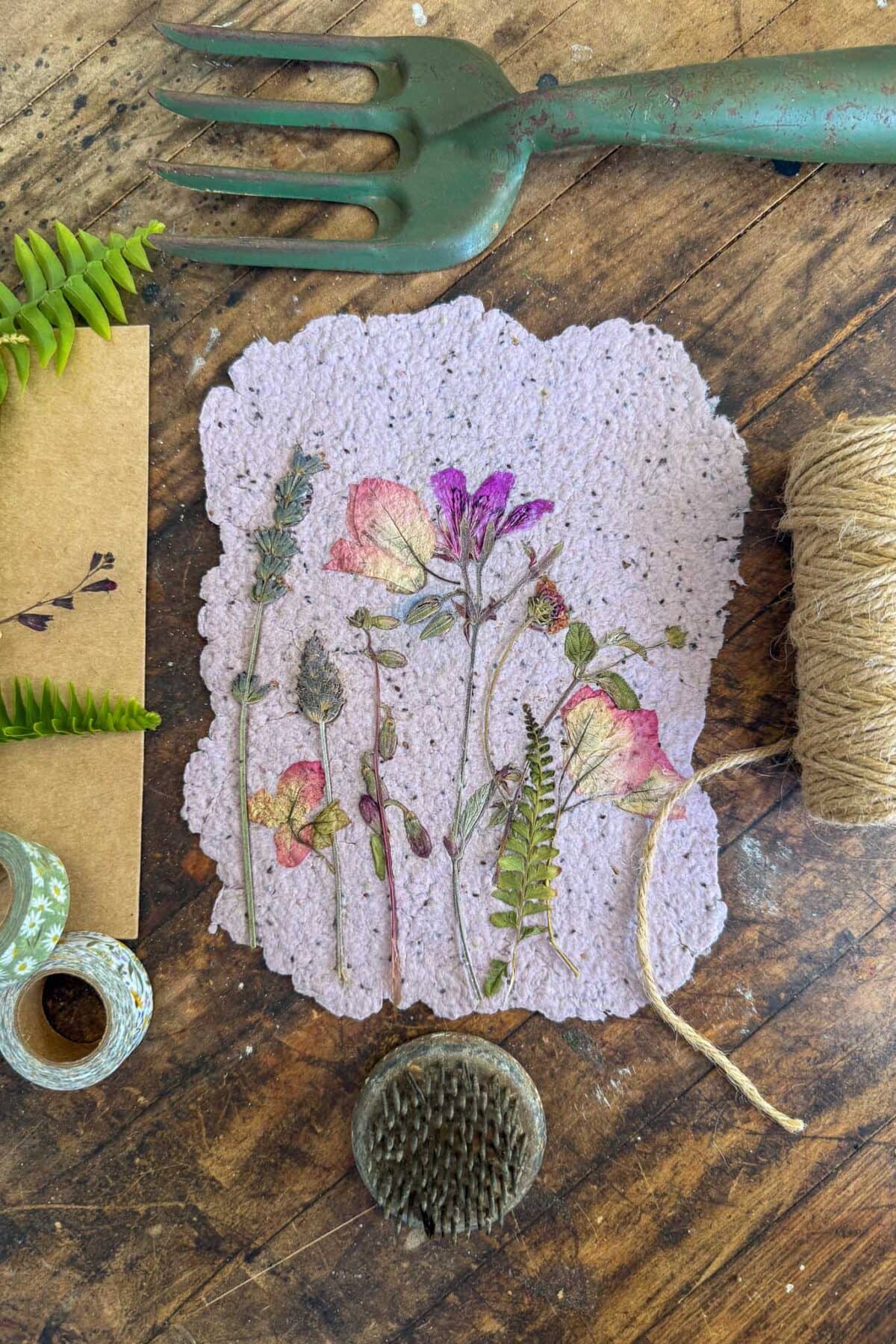 A handmade paper sheet with pressed flowers and leaves lies on a rustic wooden table, surrounded by twine, green washi tape, a metal garden fork, a floral pin holder, and an envelope with a fern leaf.
