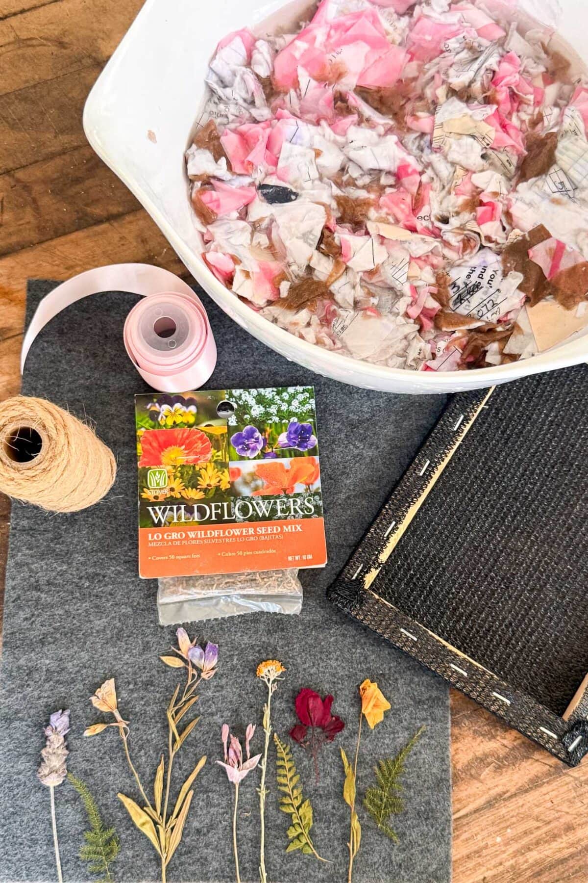 A crafting setup with a tray of wet, torn pink and white paper, a spool of twine, pink ribbon, a wildflower seed packet, a closed black notebook, and various dried flowers arranged on a felt mat.