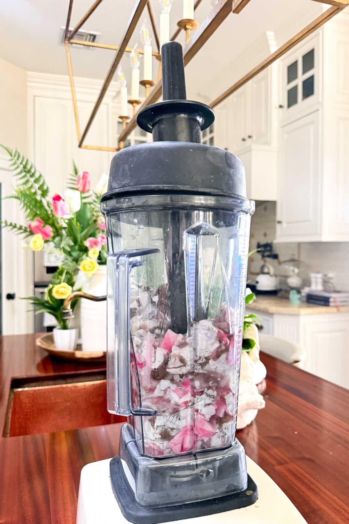 A blender filled with chopped dragon fruit and other ingredients sits on a wooden kitchen counter, with white cabinets, a light fixture, and a vase of colorful flowers in the background.