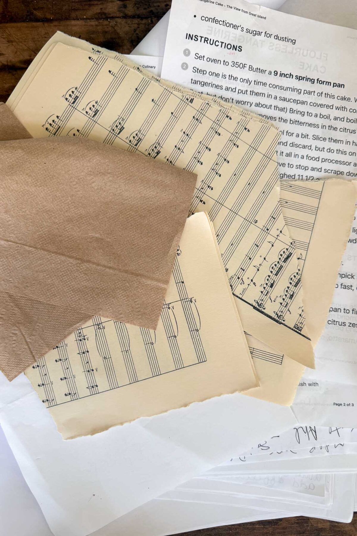 A stack of vintage sheet music partially covered by a brown napkin rests on top of printed recipe instructions, handmade cards, and handwritten notes on a wooden surface.