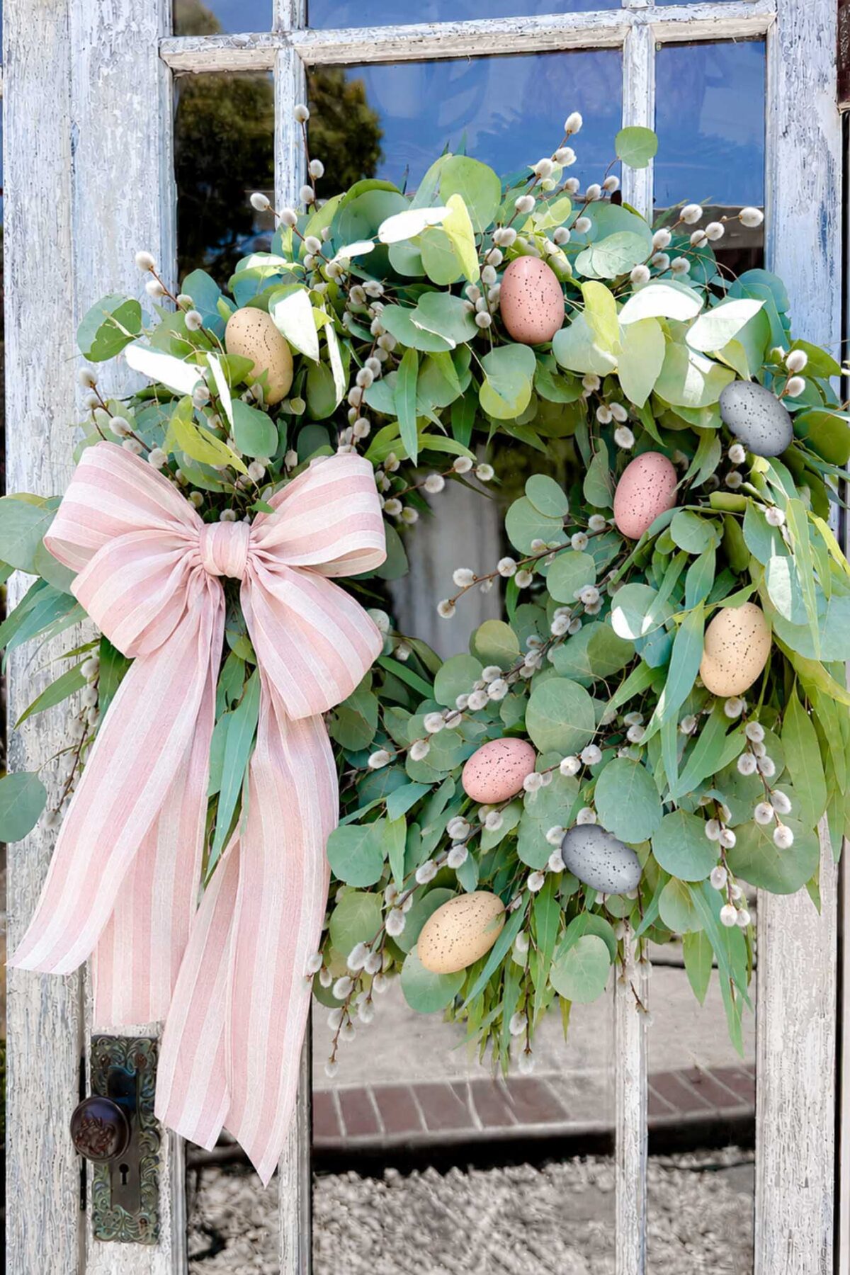 A seasonal wreath featuring green leaves, pussy willow branches, and pastel speckled eggs, with a large pink and white striped bow, hangs on a weathered glass door—perfect for those who love the look of a DIY wreath.