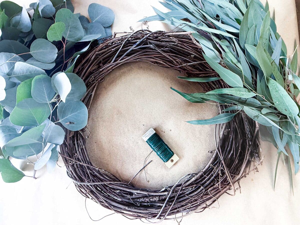 A grapevine wreath base sits on a brown background, surrounded by eucalyptus branches. A spool of green floral wire is in the center of the wreath.