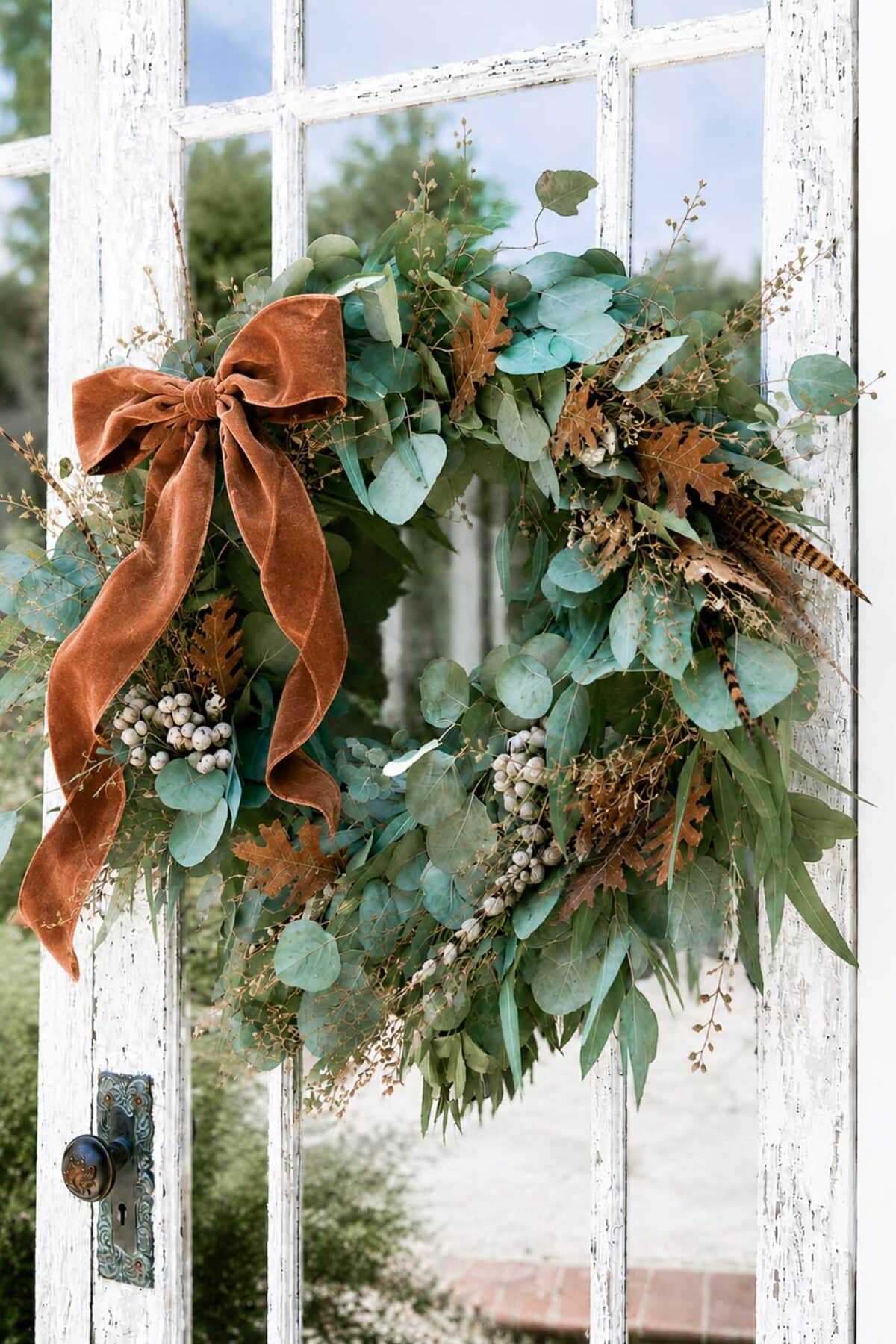 A rustic wreath of eucalyptus, dried leaves, berries, and feathers hangs on a white, distressed wooden door, adorned with a burnt orange velvet bow.
