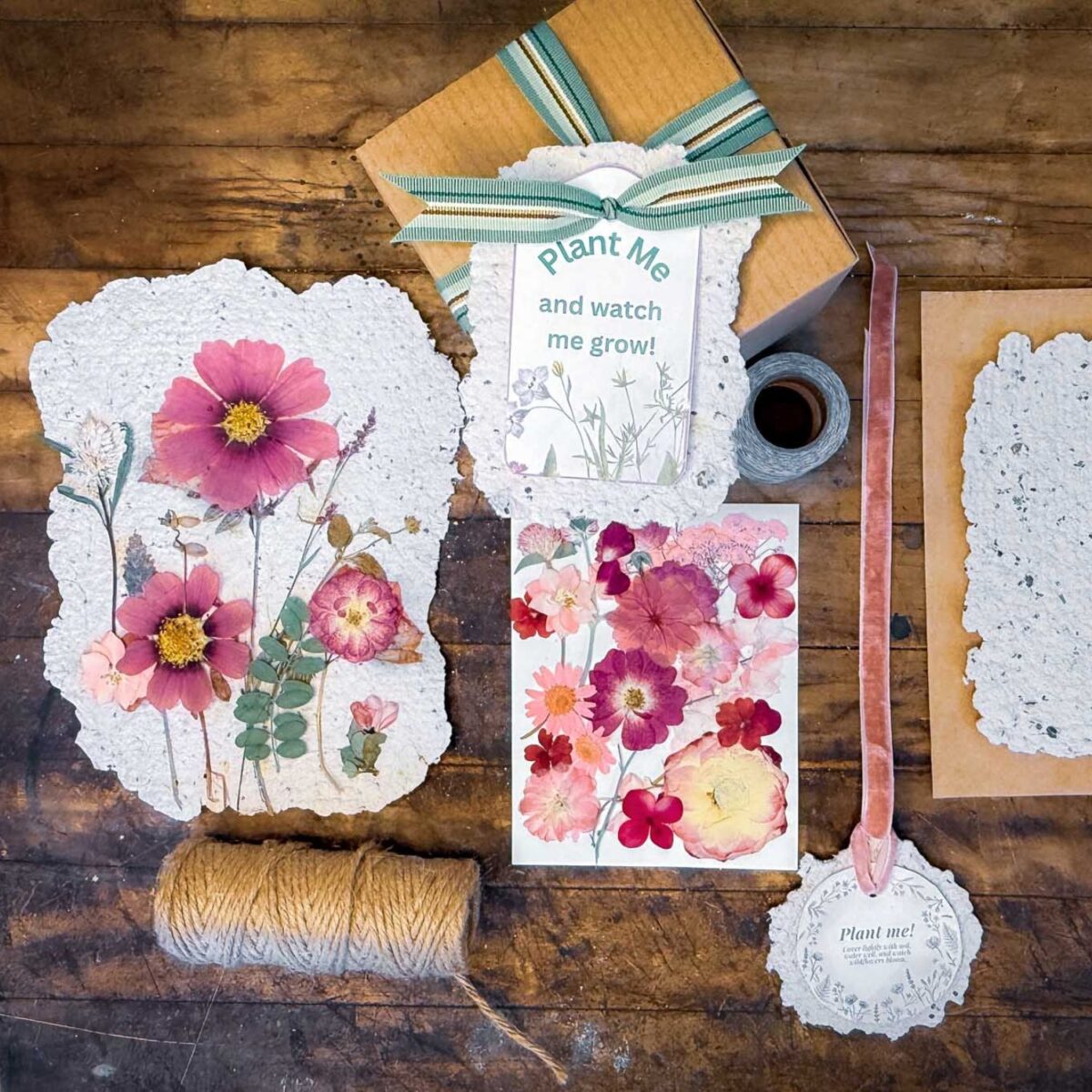 A flat lay of handmade DIY seed paper sheets with pressed flowers, a "Plant Me and watch me grow!" tag, spool of twine, ribbon, tape, and a box on a rustic wooden surface, suggesting a charming plantable paper craft kit.