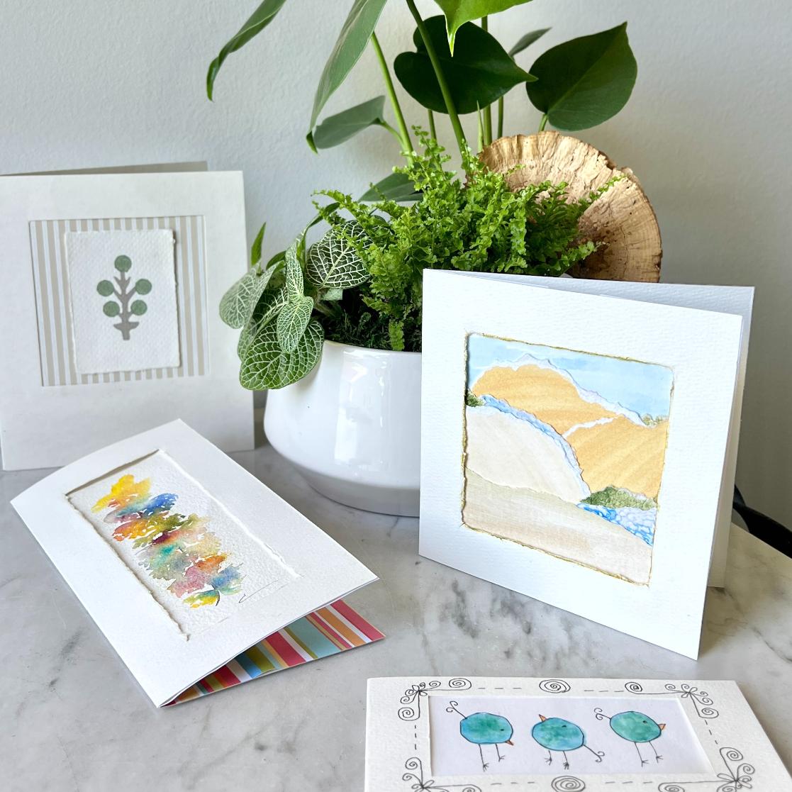 Four handmade cards crafted on DIY seed paper, featuring watercolor and ink designs, are displayed on a marble surface beside a white pot with green leafy plants. The cards showcase abstract art, trees, and plant-inspired motifs—perfect eco-friendly stationery.