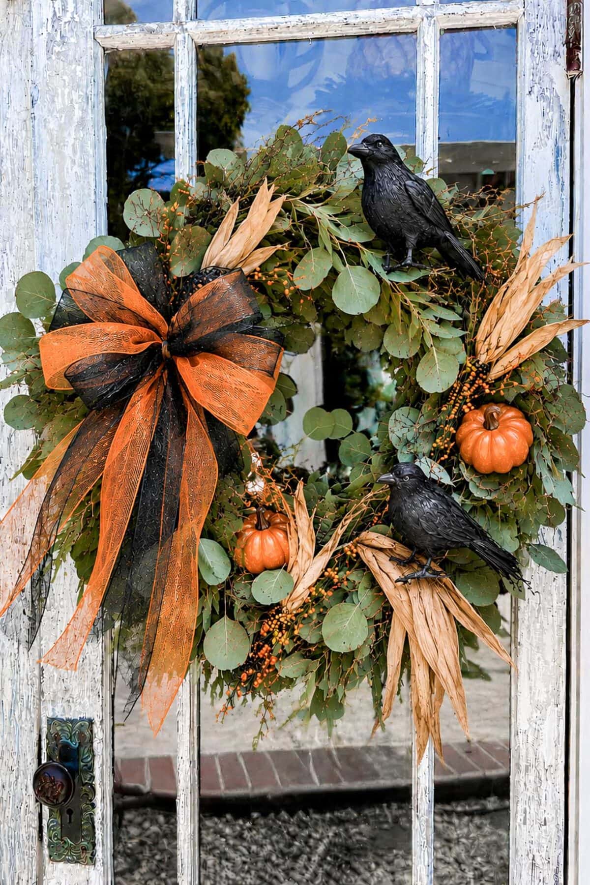 A wreath decorated with orange and black ribbon, artificial pumpkins, green leaves, and two black crows hangs on a weathered, white wooden door with glass panels.