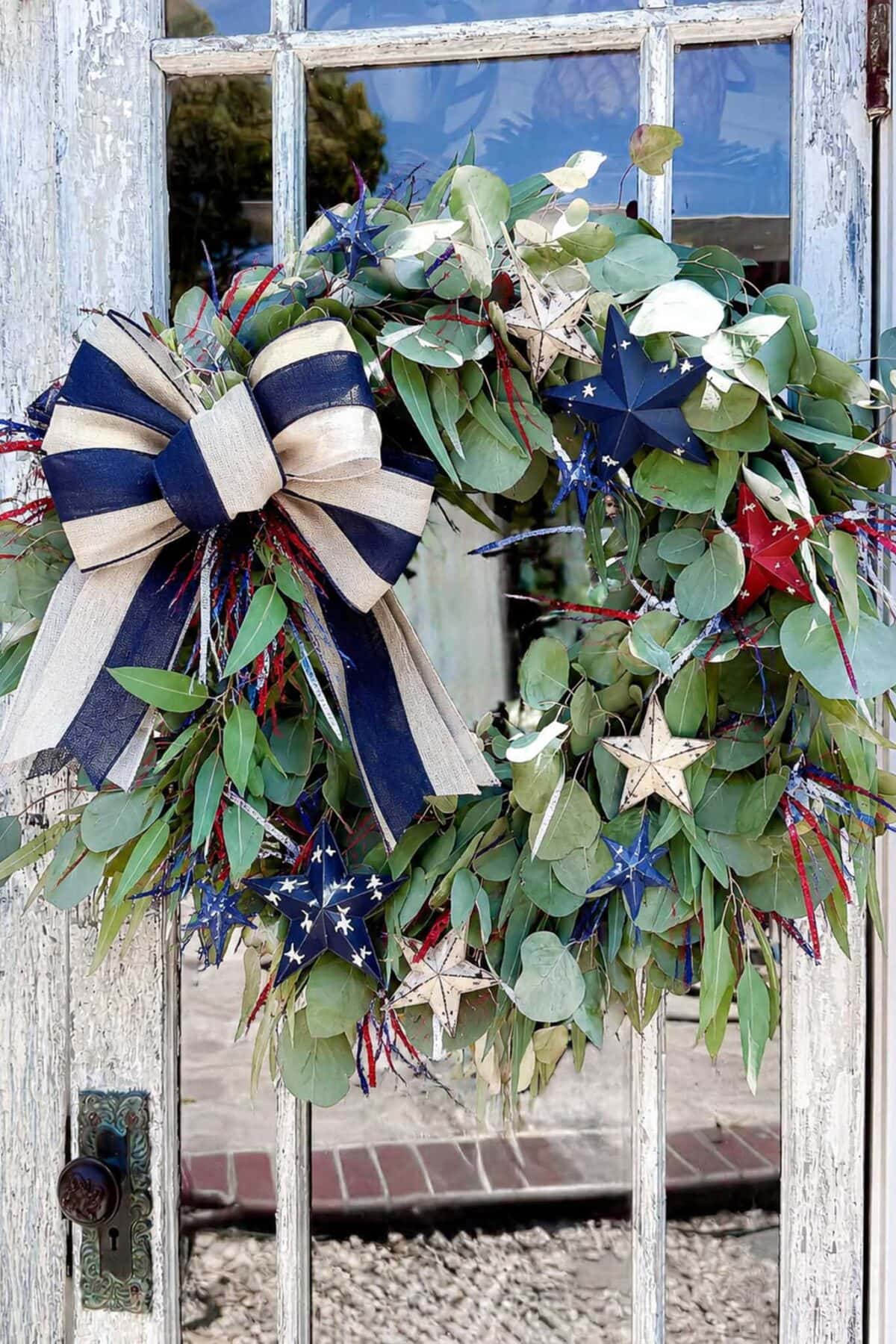 A leafy wreath decorated with red, white, and blue star ornaments, shiny tinsel, and a large striped bow hangs on a weathered glass door.